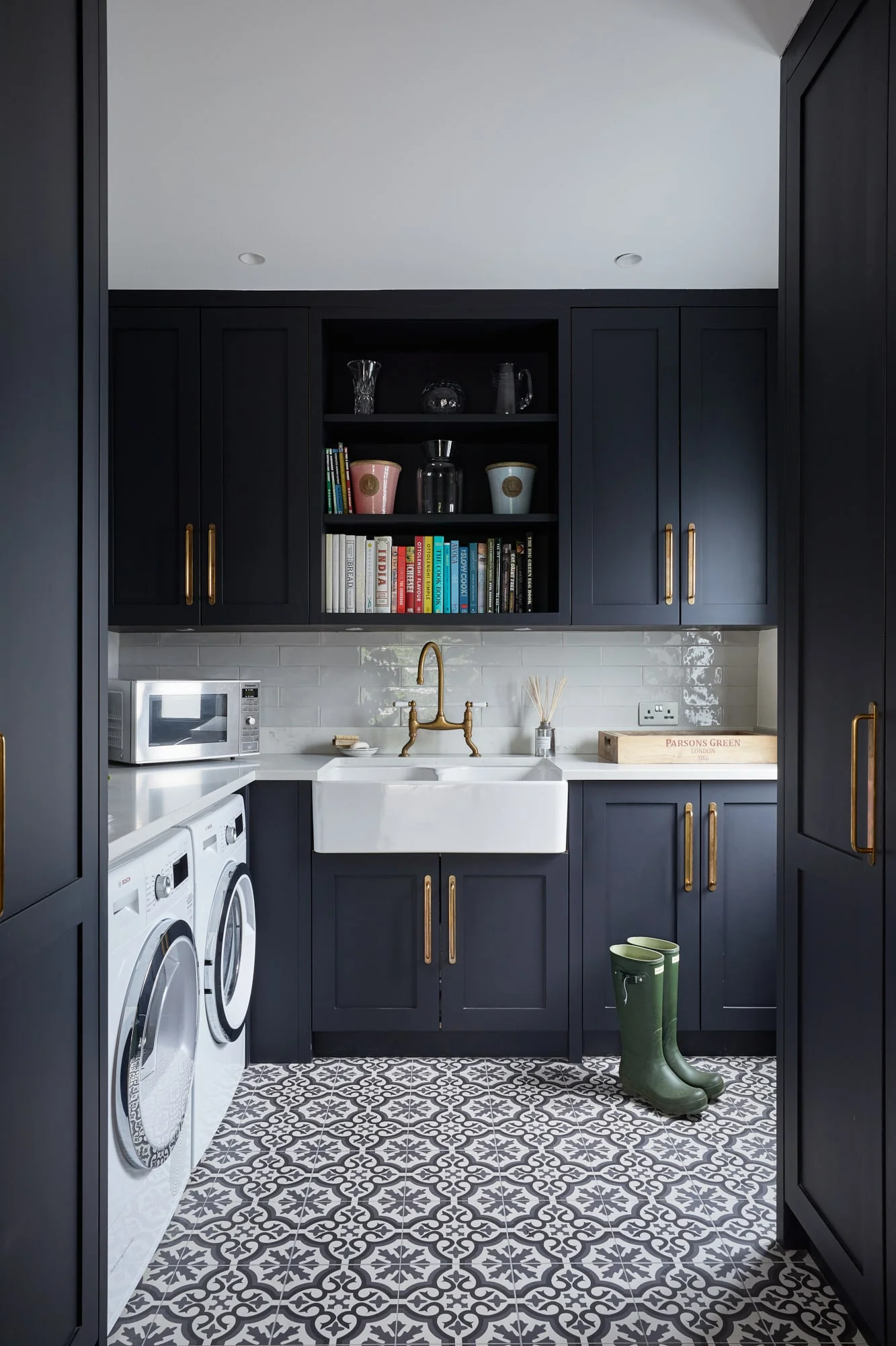 A utility and laundry room featuring dark navy cabinetry, brass hardware, patterned floor tiles, and a white Belfast sink.