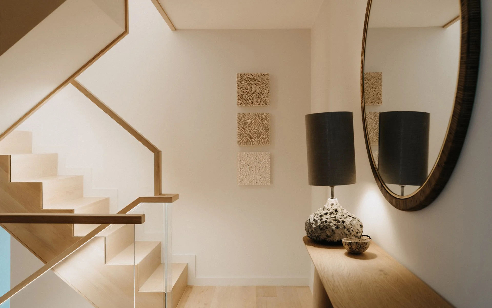 Interior stairwell with light wood treads, glass guardrail, and console table with textured lamp by MortonScarr Architects.