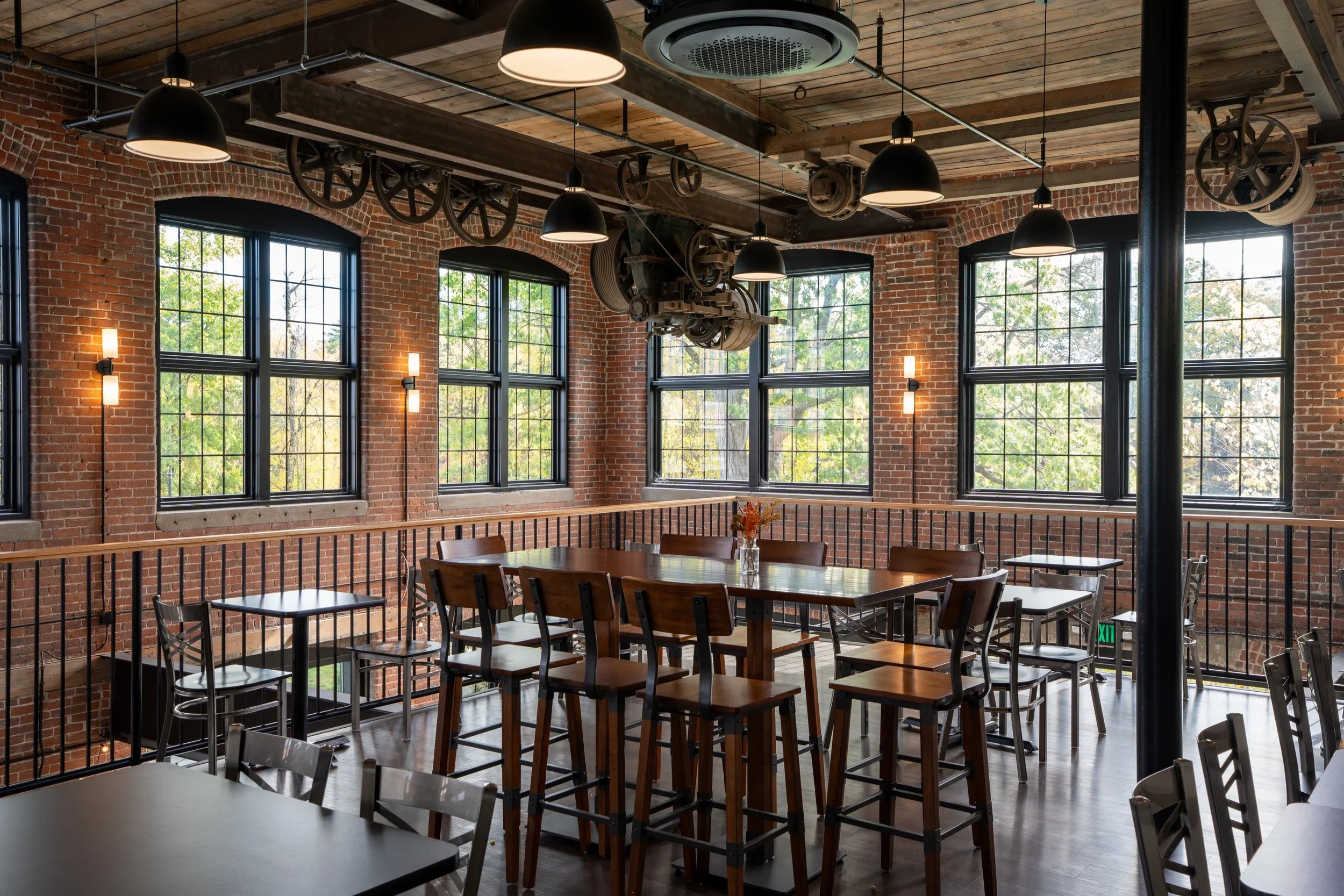 Interior of a restored industrial brick mill building with multi-pane arched windows, exposed timber ceiling joists, vintage machinery gearing on walls, and dining furniture by Bonham & Douglas Architects.