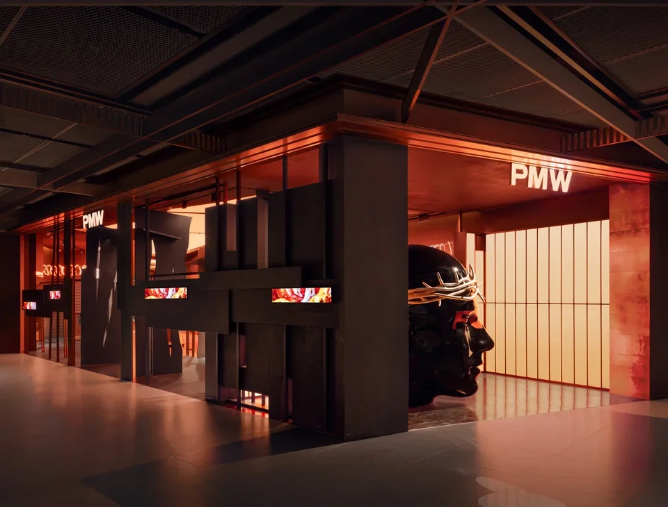 Interior retail storefront with dark metallic panels, glowing PMW signage, and a large dark sculptural head wearing a crown.
