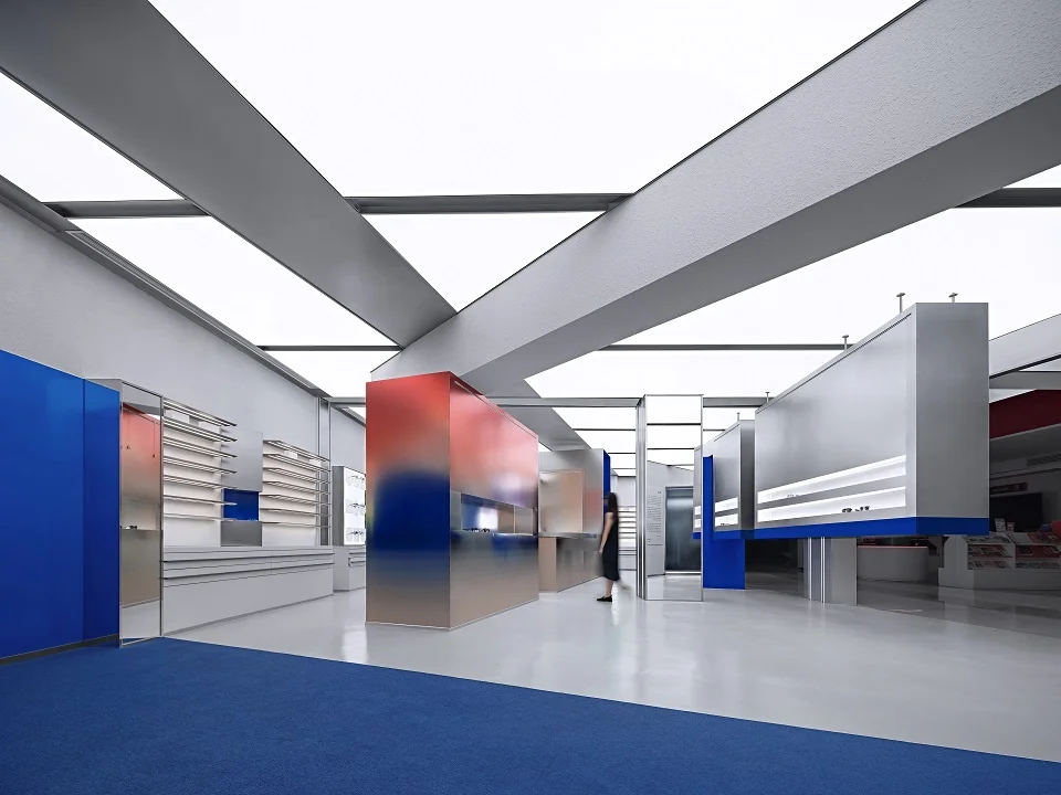 Interior of Asian-Crew concept store by 1-idesign featuring massive diagonal structural roof beams, blue carpet, and gradient metallic display volumes.
