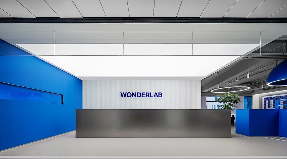 Modern corporate reception area with a metallic desk, luminous ceiling panel, and blue accent walls at Wonderlab Headquarters by 一乘建筑.