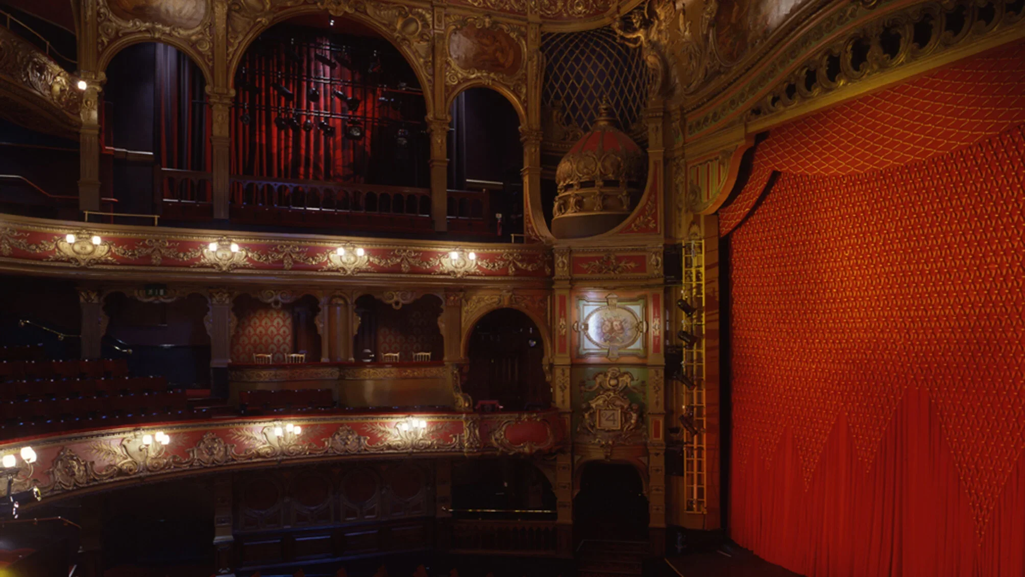 Interior view of historic theater auditorium with ornate gilt balconies and a large red velvet stage curtain by Tim Ronalds Architects.