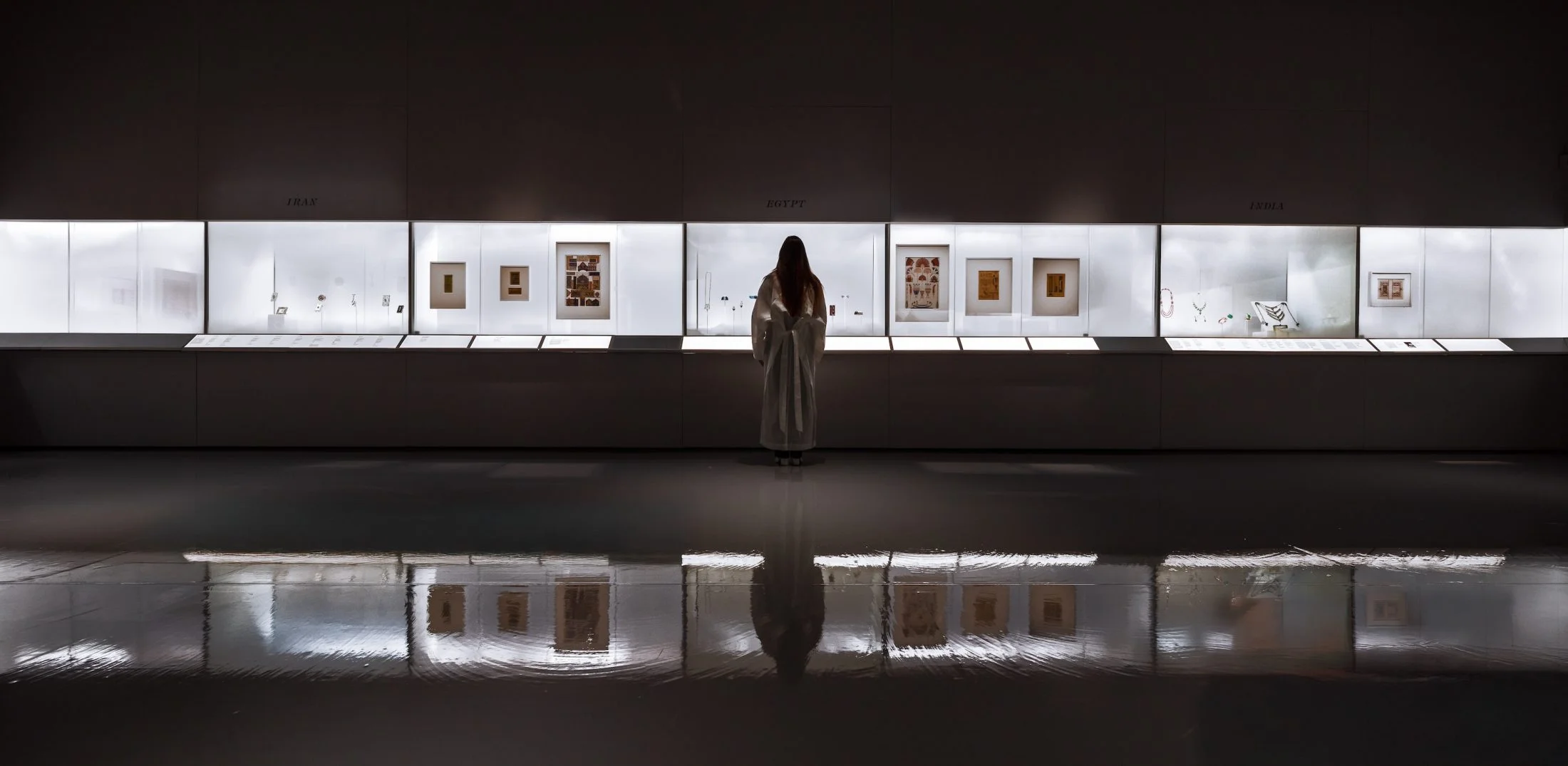 Exhibition interior with back-lit display vitrines and a dark reflective floor by Asif Khan Studio.