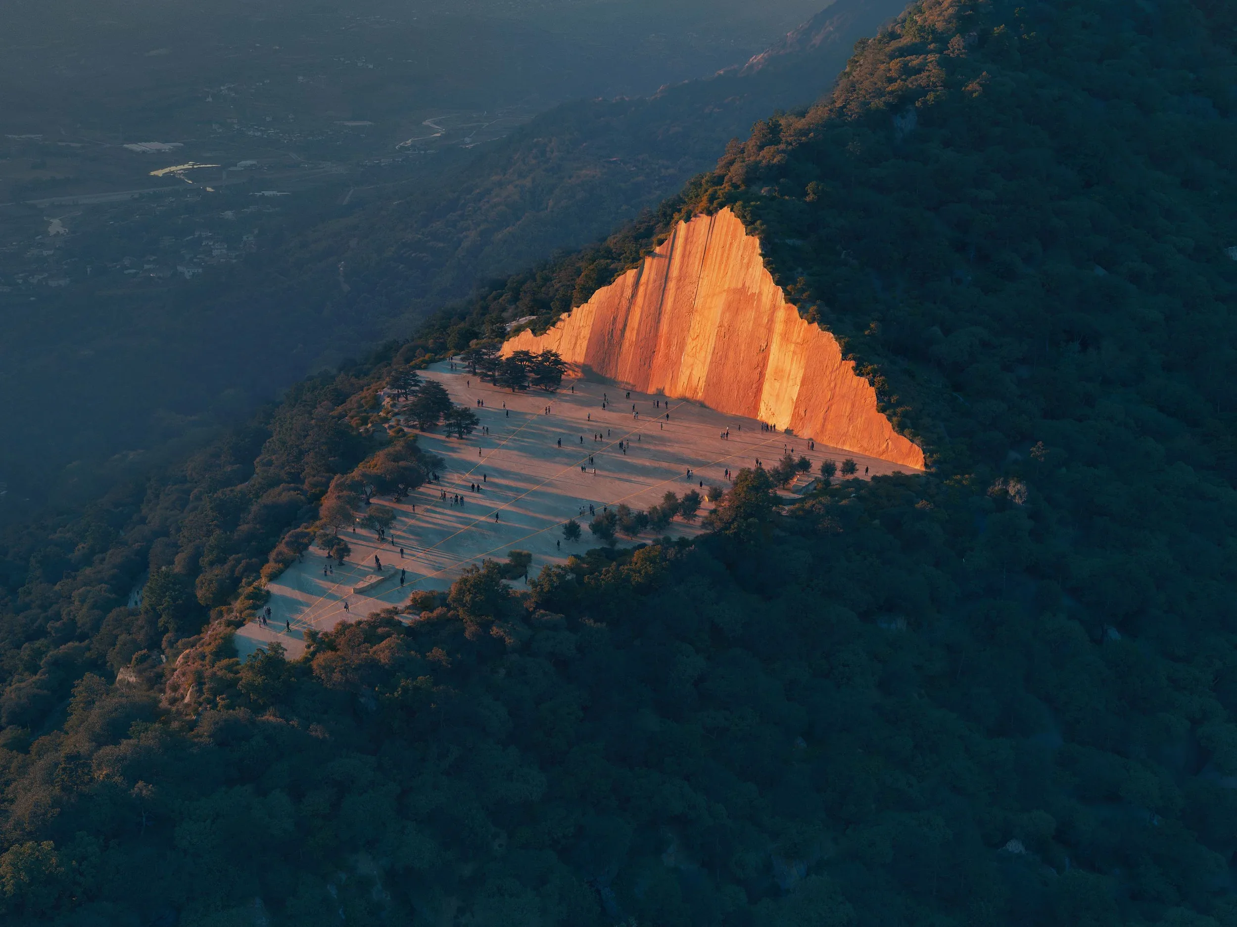 Aerial view of Faith Park by AL_A showing a large triangular plaza carved out against a massive vertical stone cliff bathed in sunset light.