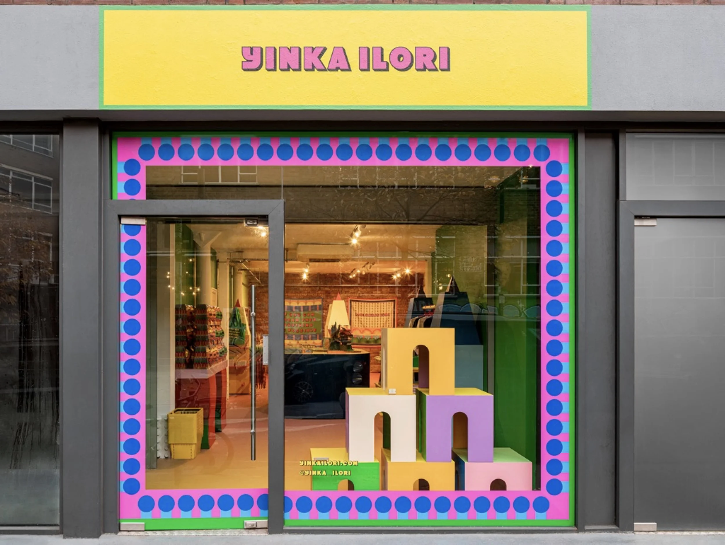 Storefront facade with bright yellow sign reading YINKA ILORI, vibrant pink-and-blue dotted window graphics, and colorful display blocks by Yinka Ilori Studio.