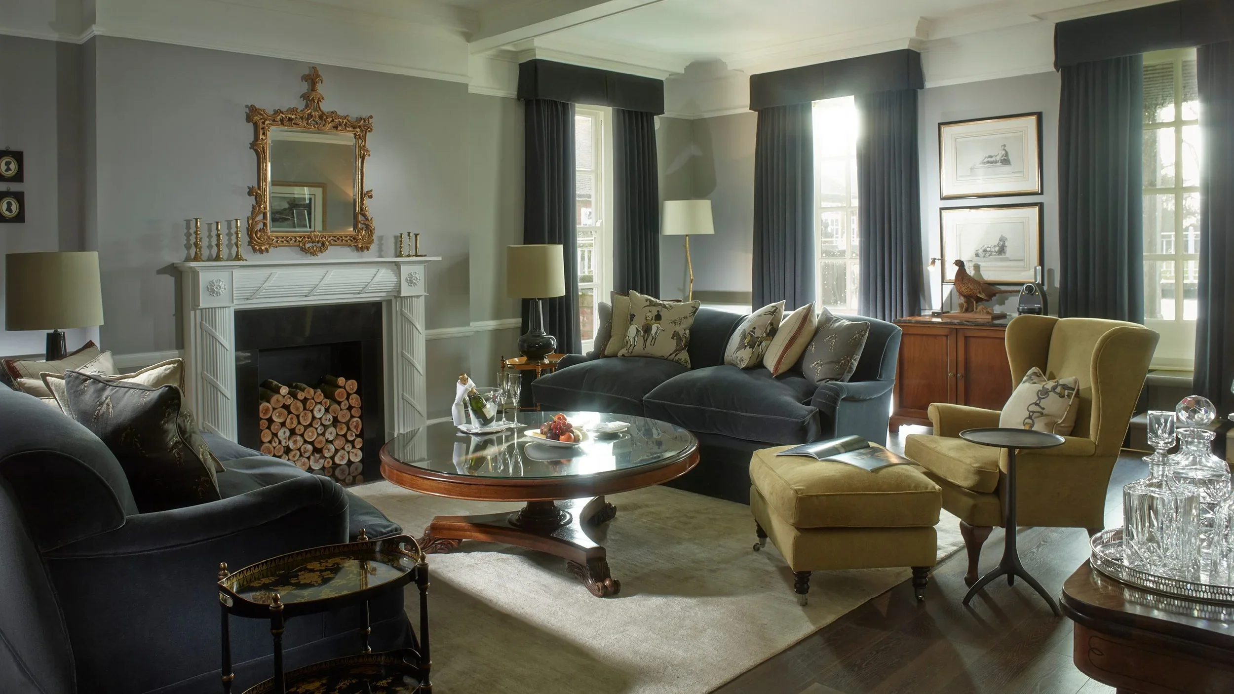 Traditional hotel drawing room with dark charcoal sofas, mustard armchairs, and carved white fireplace by Anita Rosato.