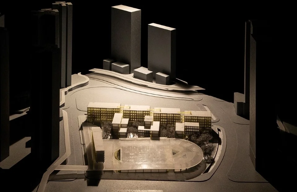 Physical architectural study model of a school campus with low-rise bar buildings and an oval running track, set among high-rise towers in a dark room.