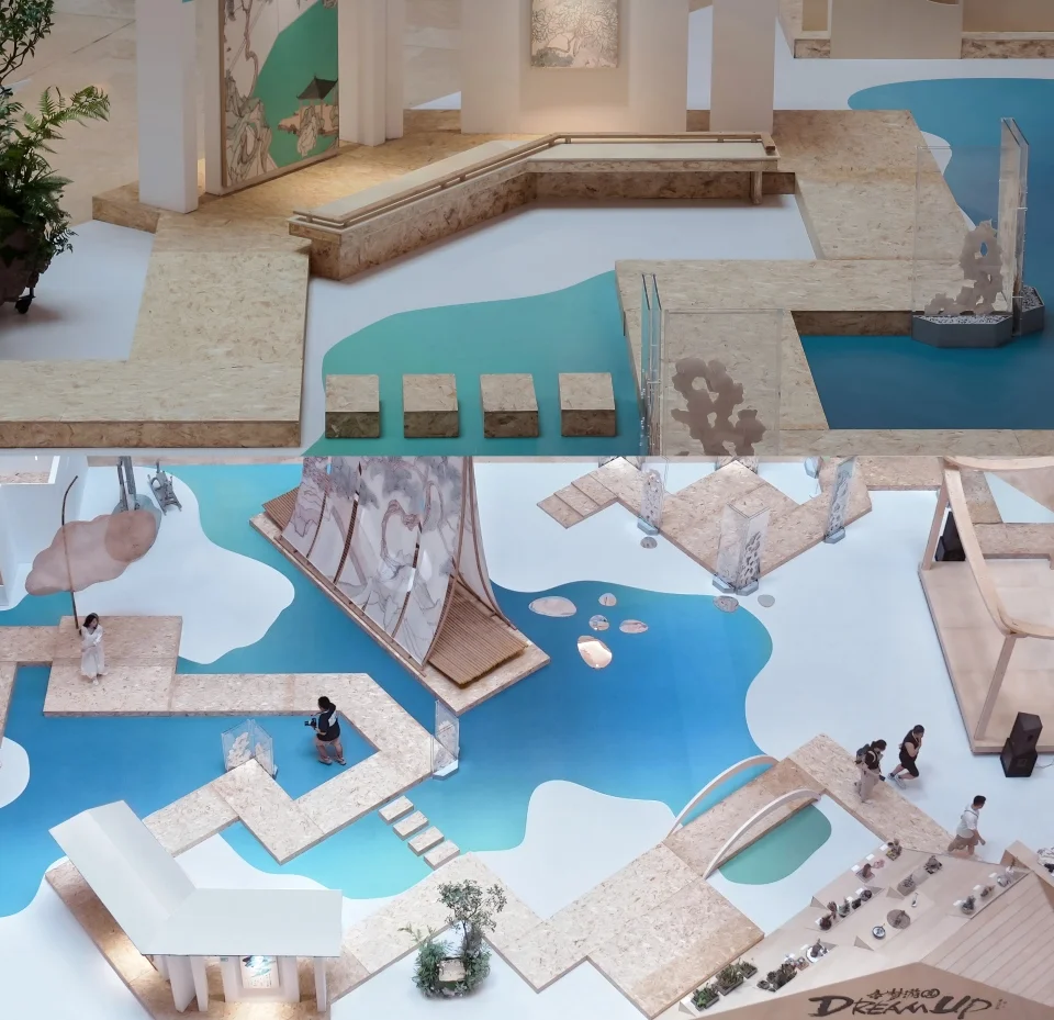 High-angle split view of an exhibition installation by 木月建筑设计事务所 with raised OSB boardwalks winding over blue painted floor graphics.