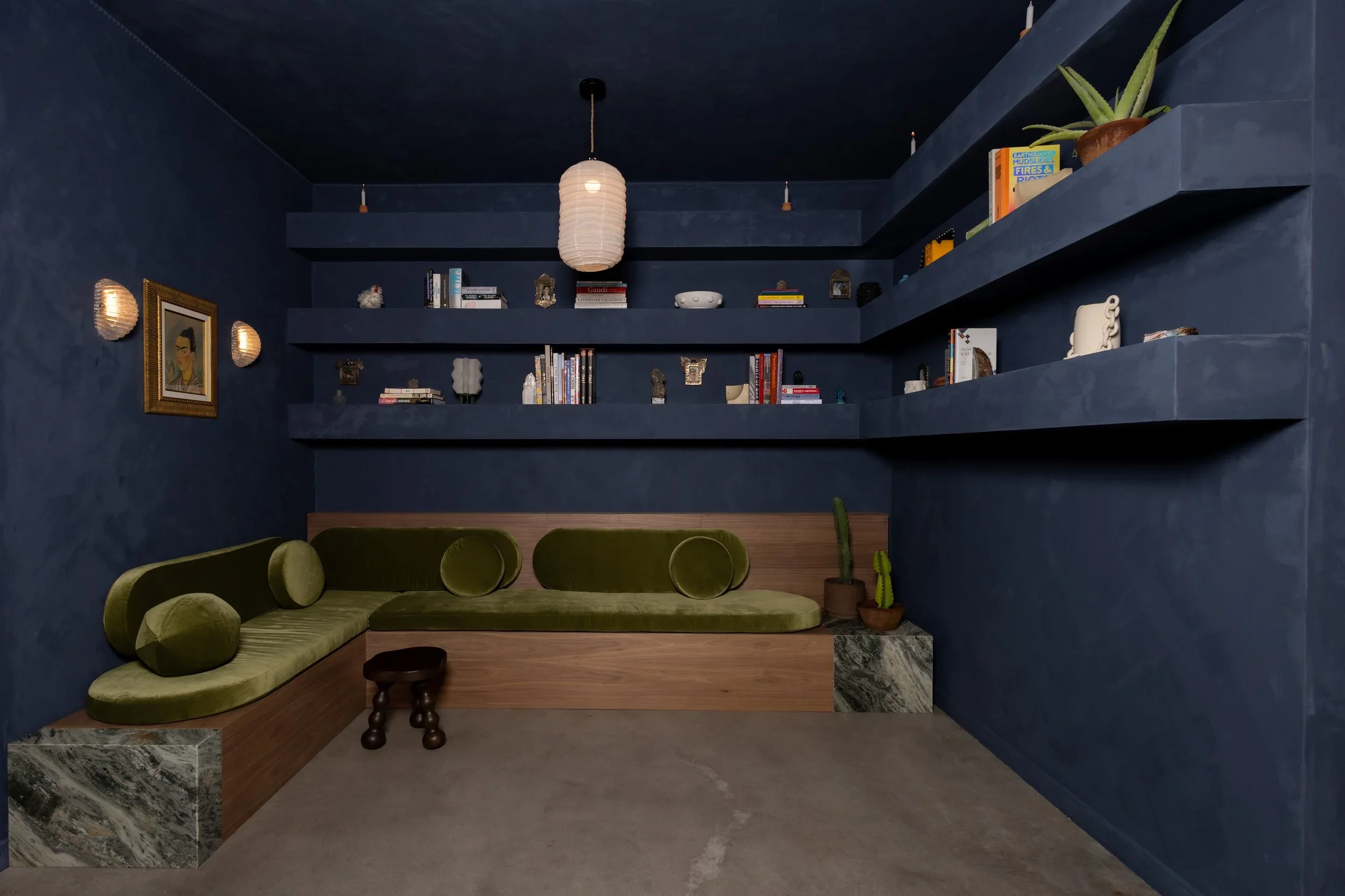 Dark blue interior lounge with built-in wooden banquette seating upholstered in green velvet, flanked by marble plinths and wrap-around floating shelves.