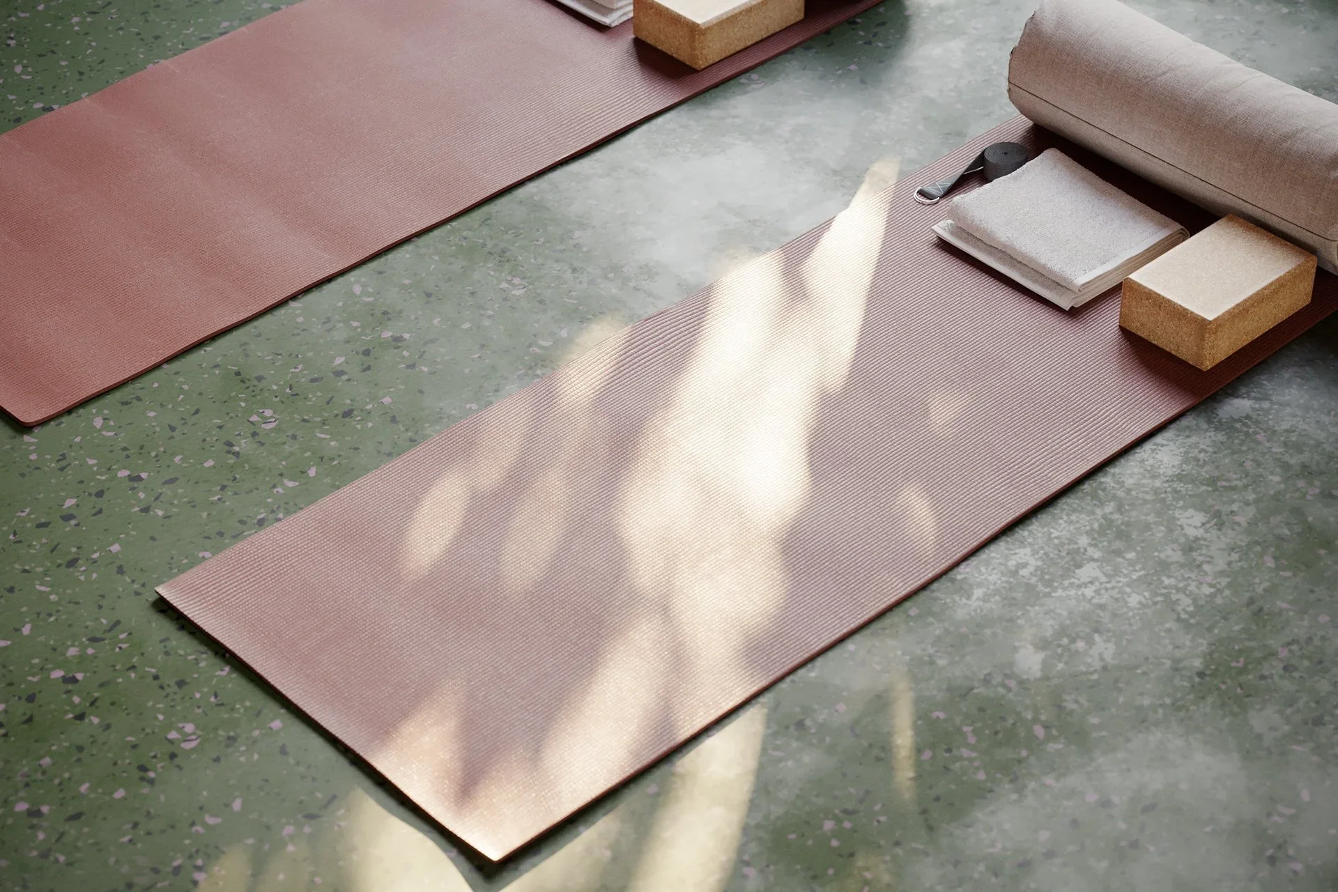 Close-up interior perspective of pink yoga mats and cork blocks on a green terrazzo floor, with dappled sunlight streaming across by Narrativ.