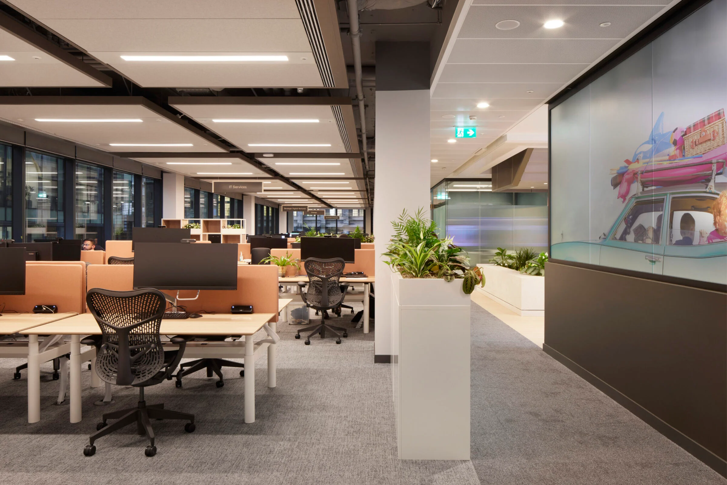 Open-plan corporate office interior by The Furniture Practice featuring modular desks, acoustic ceiling baffles, and integrated planters.