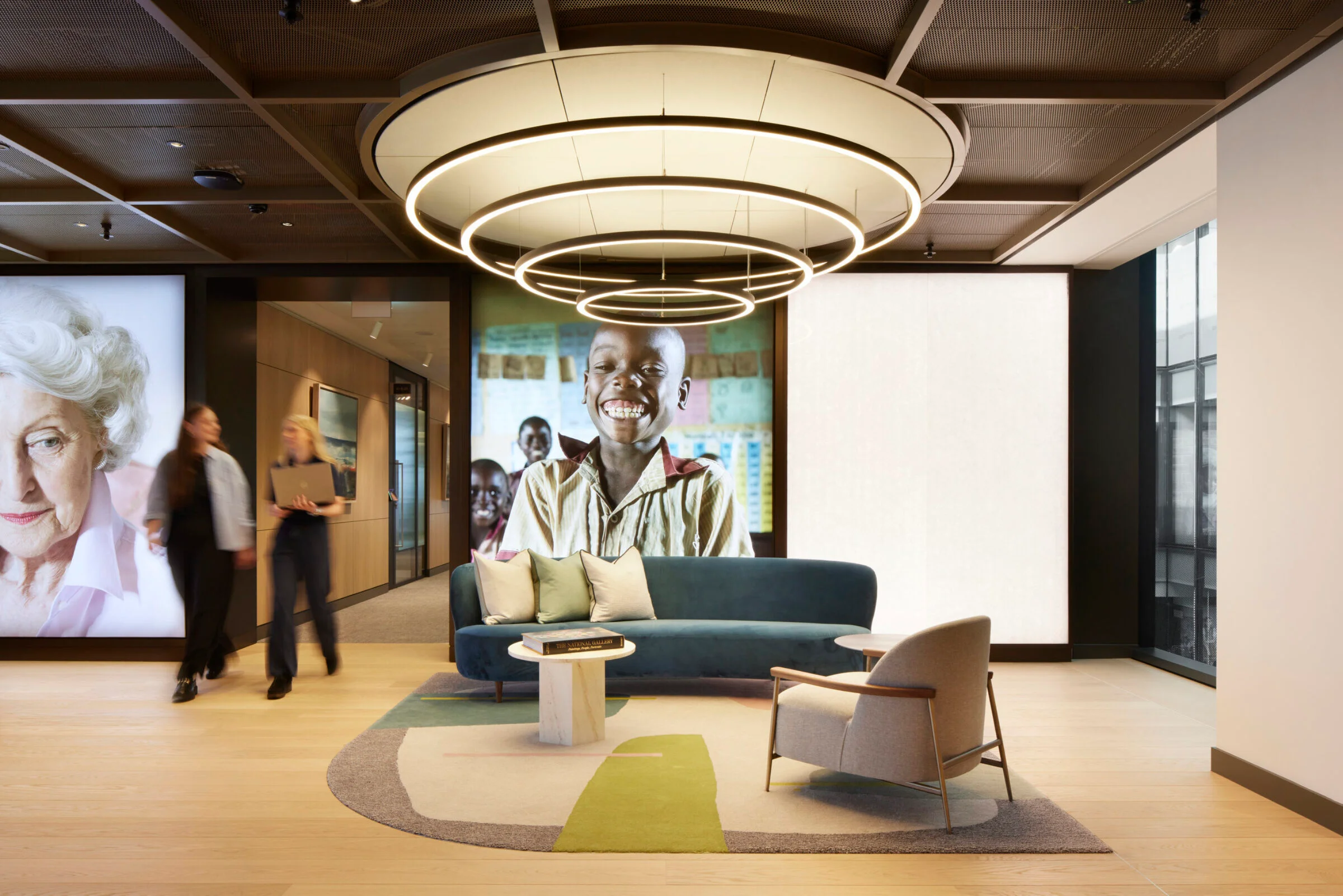 Commercial reception lounge with a large ring pendant light fixture, teal sofa, and graphic backlit wall panels by The Furniture Practice.