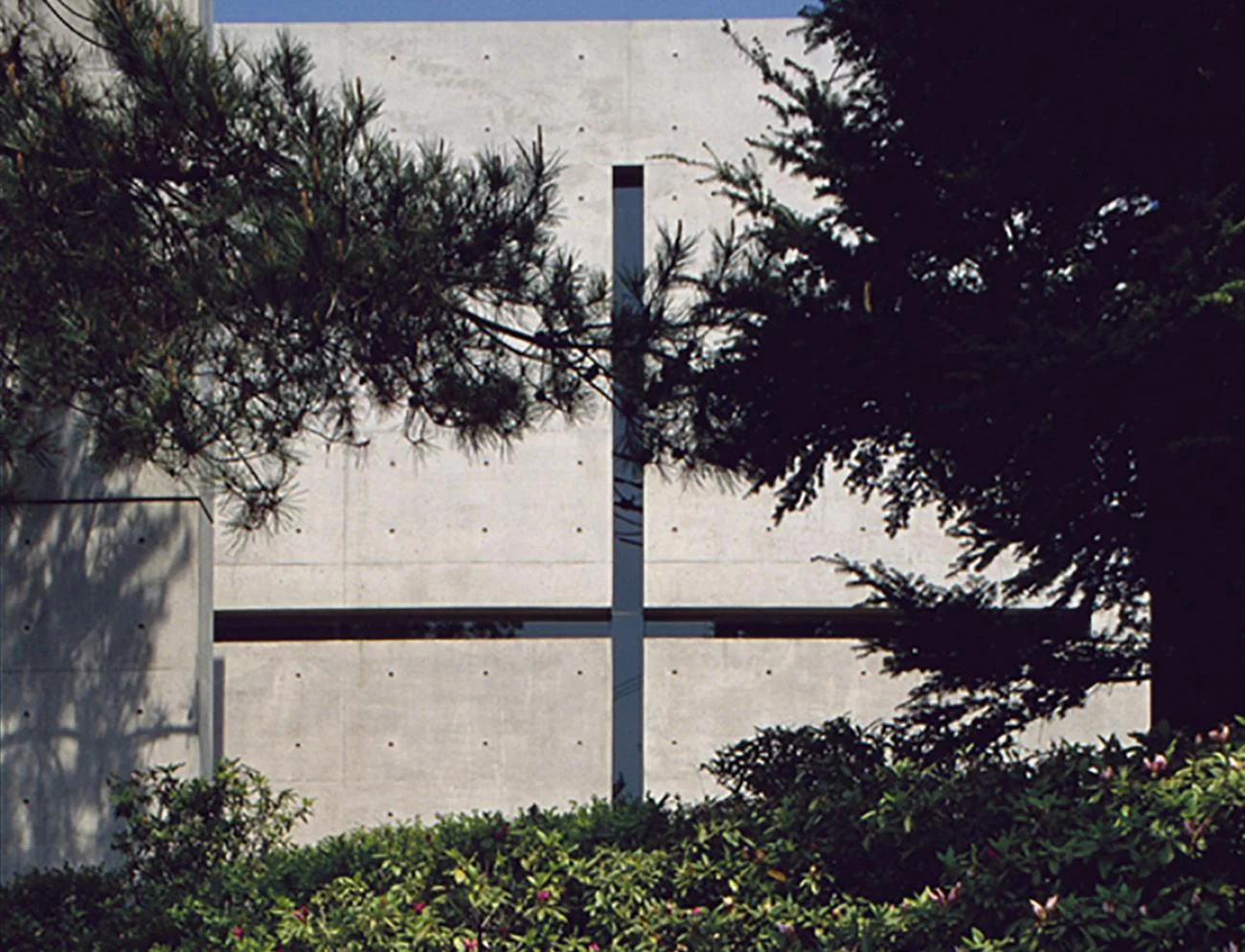 The concrete exterior facade of Tadao Ando's Church of the Light, featuring a cross-shaped cutout framed by trees.