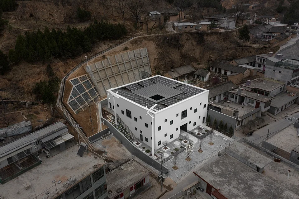 High-angle aerial view of a white courtyard building with scattered small windows nestled into a hillside village.