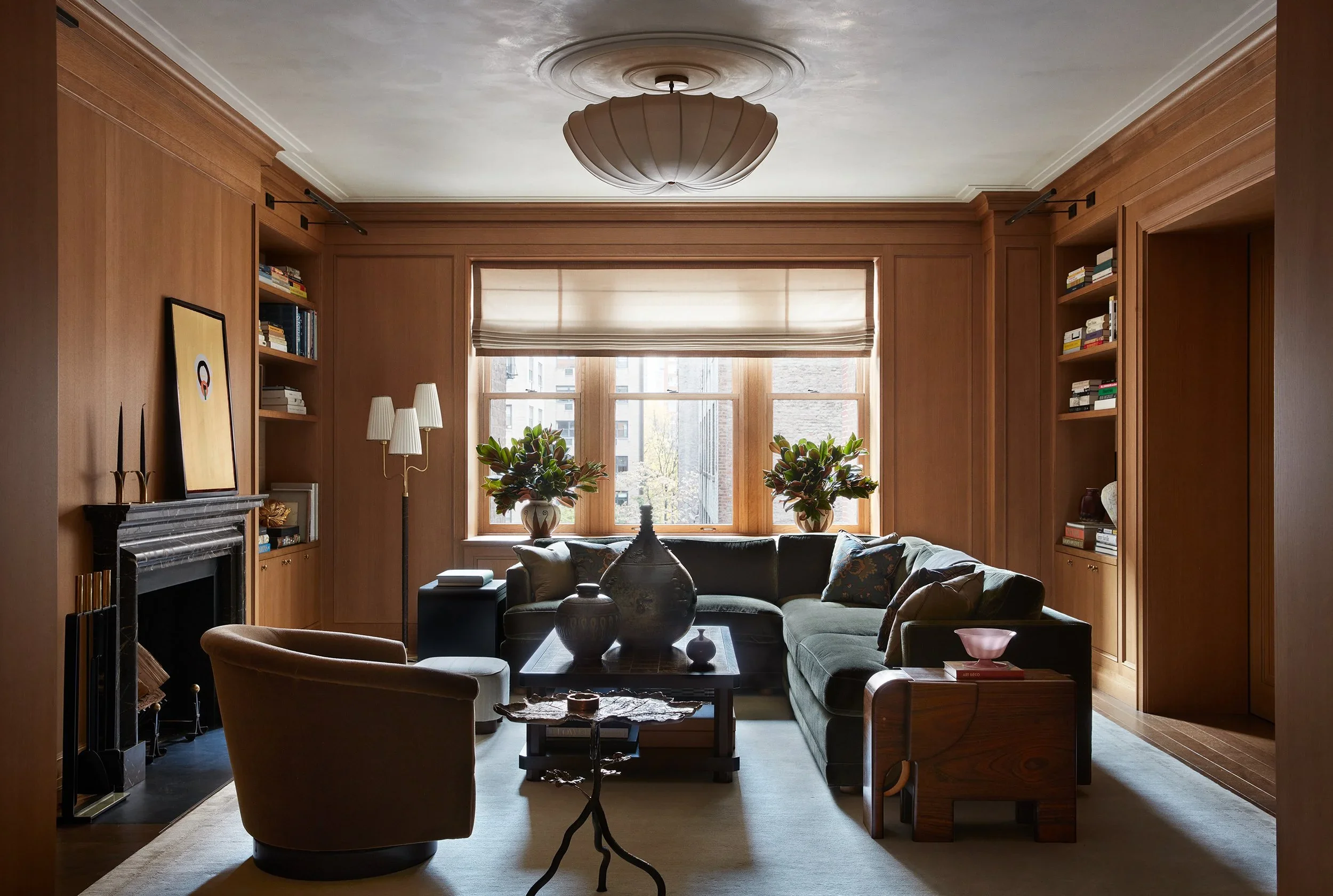 Wood-paneled living room featuring built-in bookshelves, a dark marble fireplace, and a deep green sectional sofa by Gachot Studios.