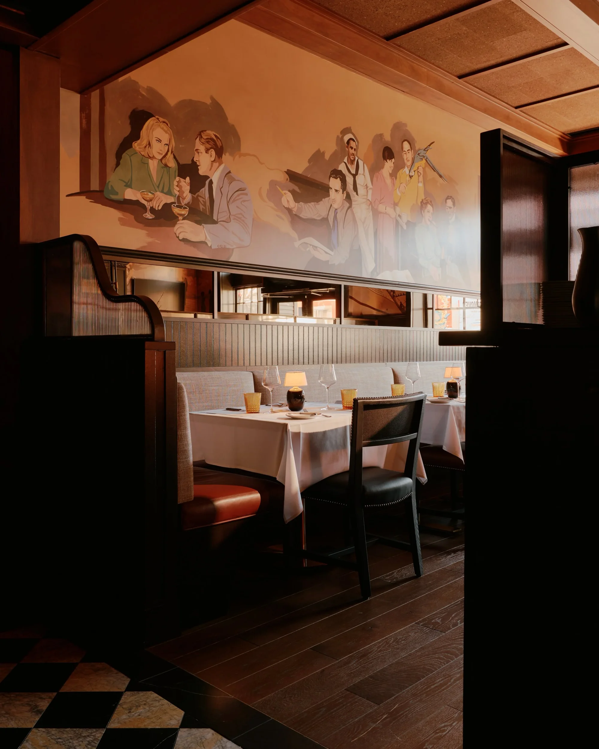 Interior dining booth with upholstered banquette, white tablecloth, dark wood partitions, and large wall mural by Gachot Studios.
