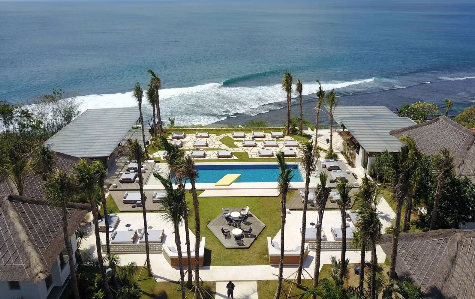 Aerial view of a coastal resort pool deck flanked by palm trees and outdoor lounges overlooking the ocean.