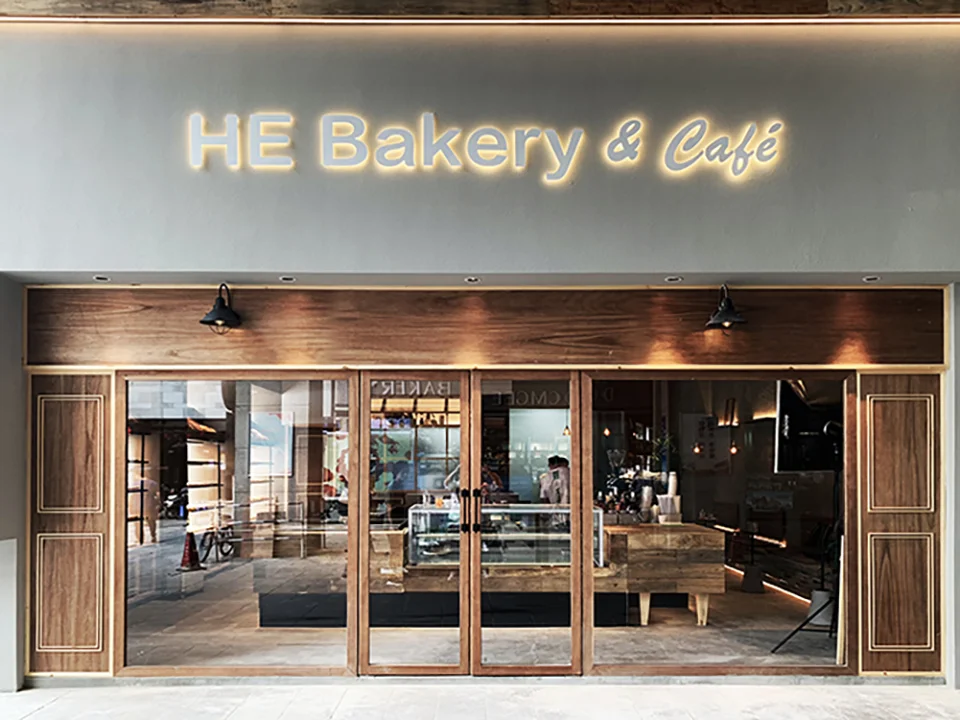 Exterior facade of HE Bakery & Cafe with wood-framed glass storefront, warm lighting, and illuminated signage by 温度ONDO空间设计.