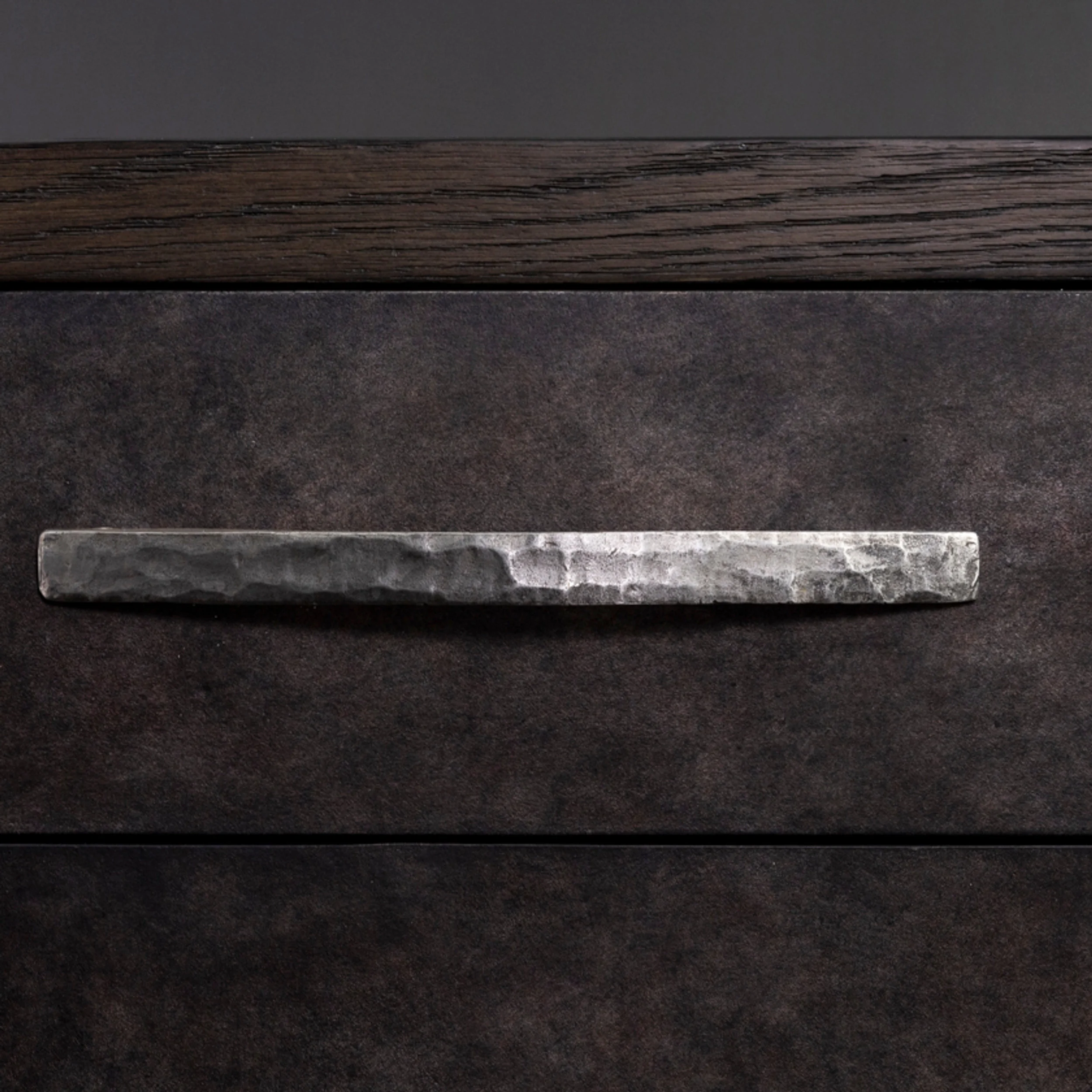 Close-up of a hammered metal drawer pull set against dark textured cabinetry by Van Thiel & Co.