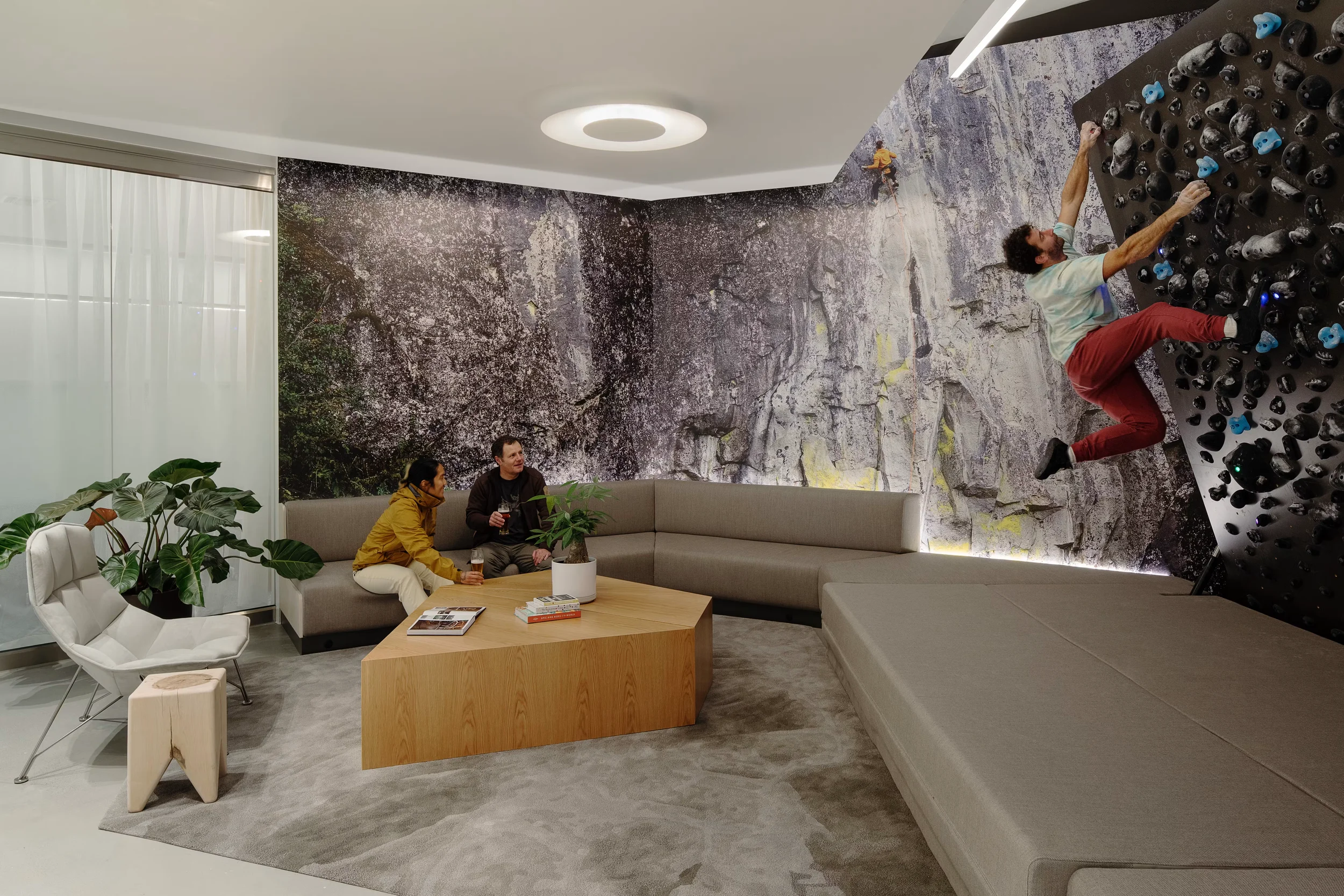 Interior office lounge by Skylab Architecture featuring a custom climbing wall, modular grey sofa, angular wood coffee table, and large rock-face wall graphic.