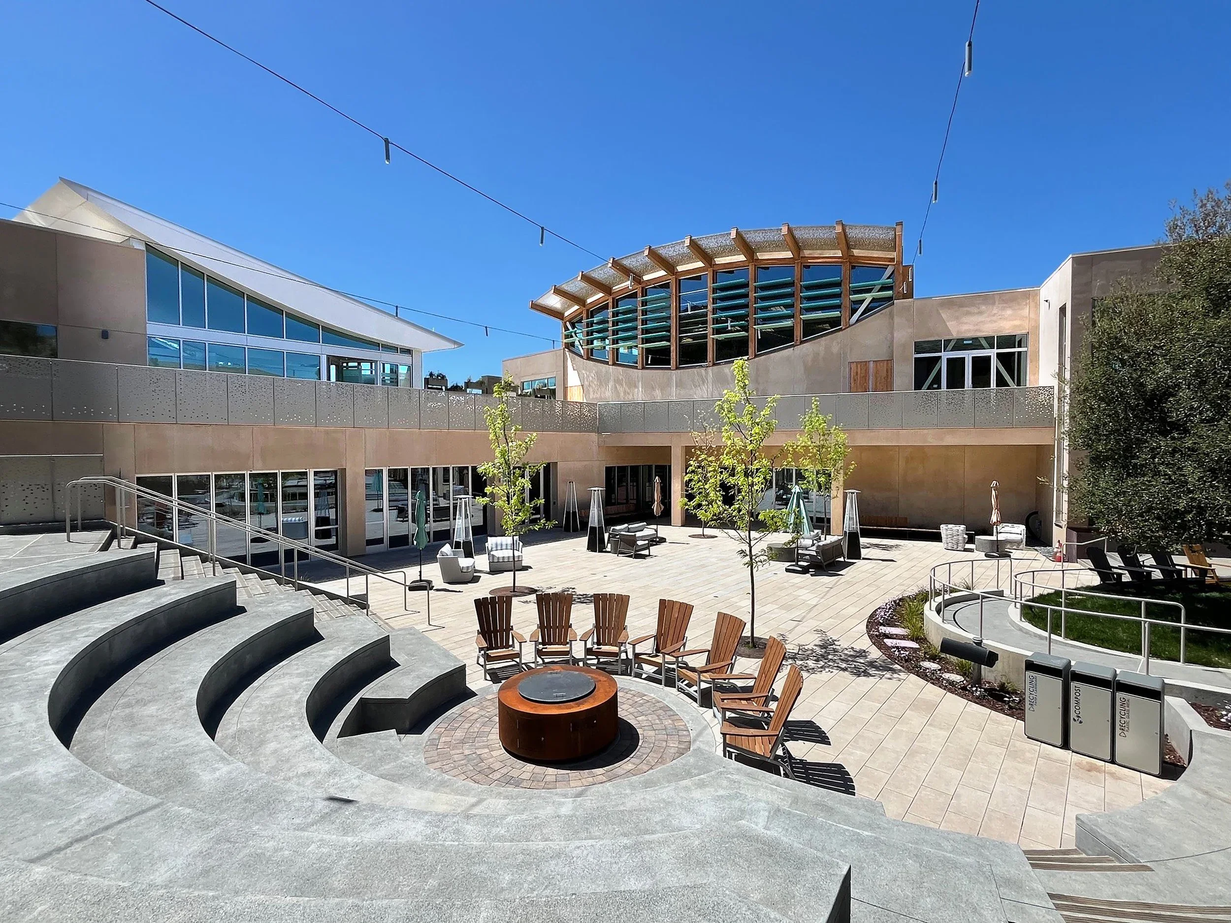 An outdoor courtyard with tiered concrete seating, a central fire pit, and curved architectural facades by Herman Coliver Locus Architecture.