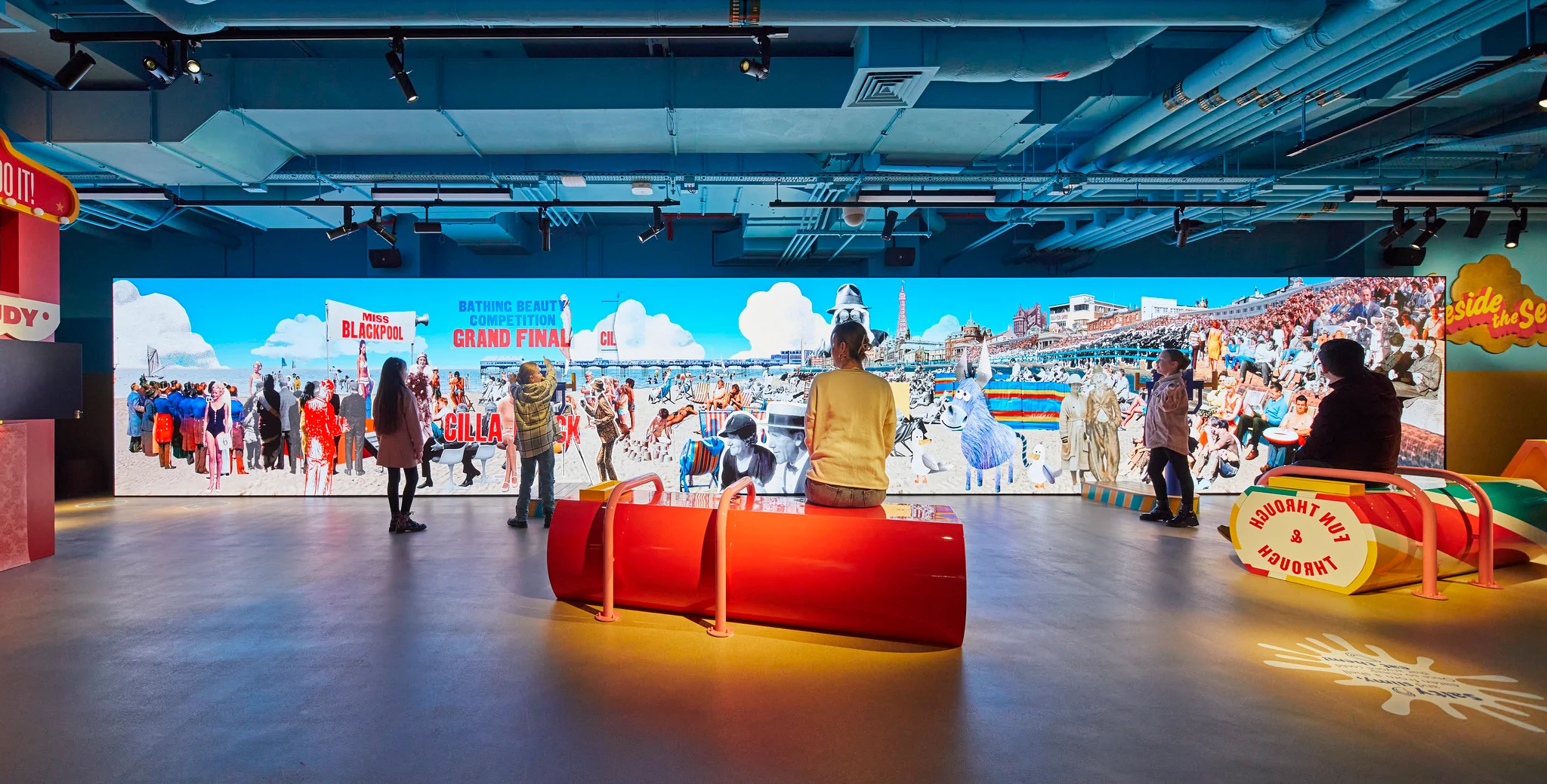 Exhibition interior by Casson Mann featuring a massive curved digital display wall and custom red rock candy seating benches.