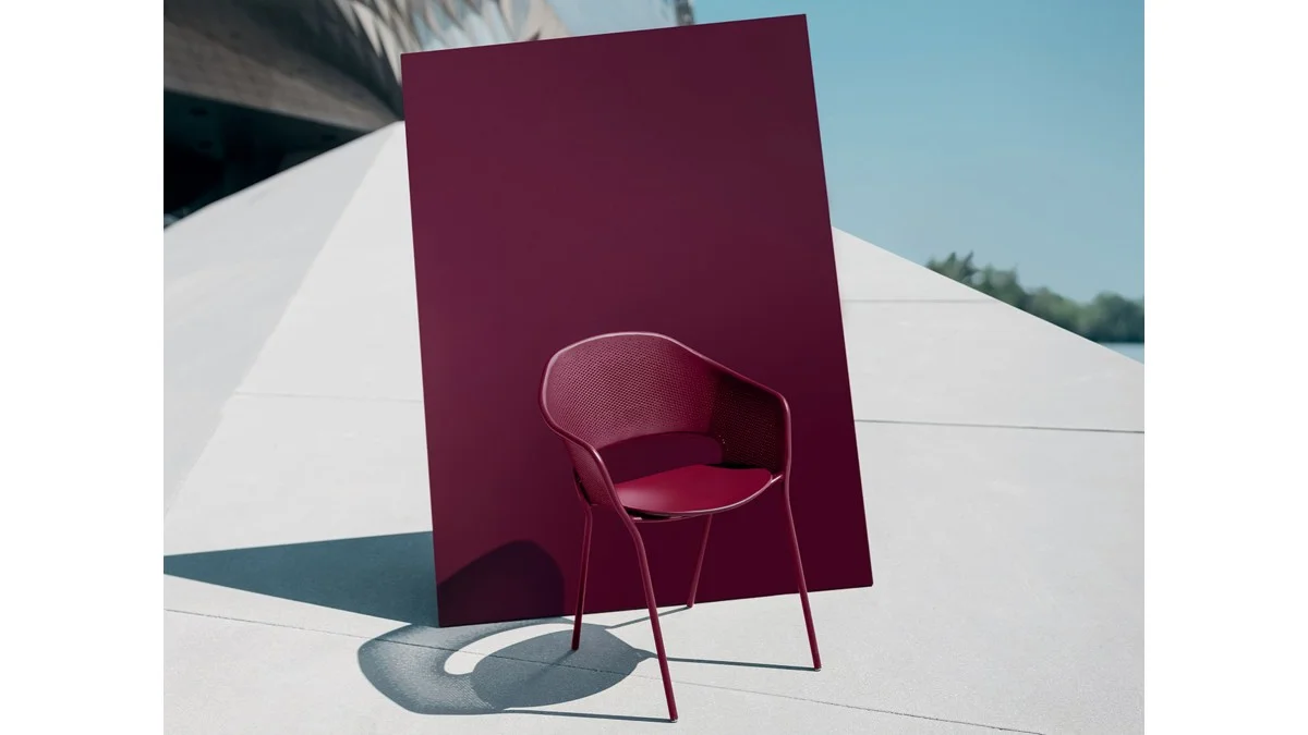 Outdoor stacking armchair in burgundy metal set against a matching backdrop on concrete paving.