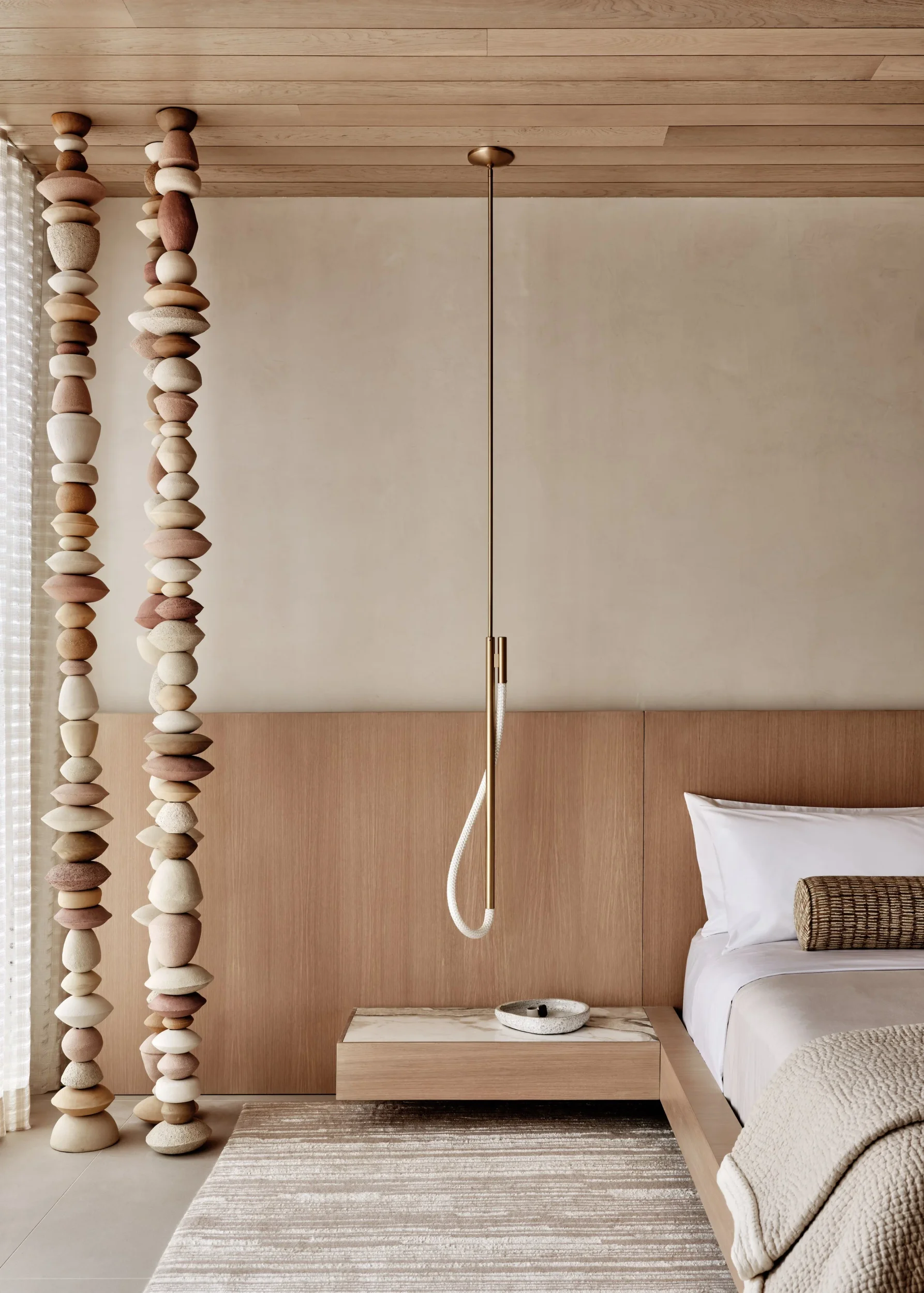 Close-up interior bedroom view featuring stacked stone columns, wood headboard, and minimalist brass pendant light by Chango & Co.
