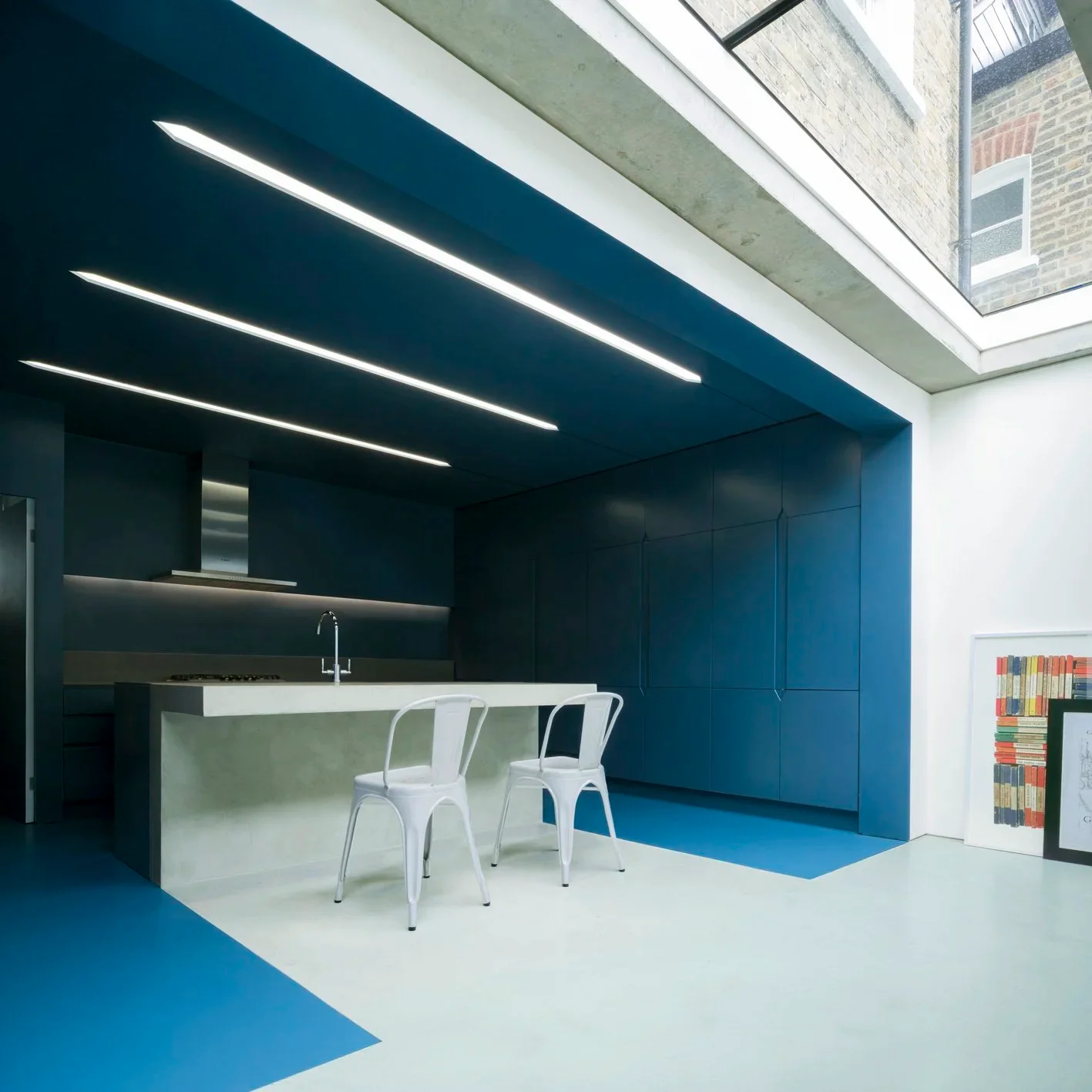 Modern open kitchen with blue cabinetry and ceiling slots, lit by a side skylight by Bureau de Change Architects.