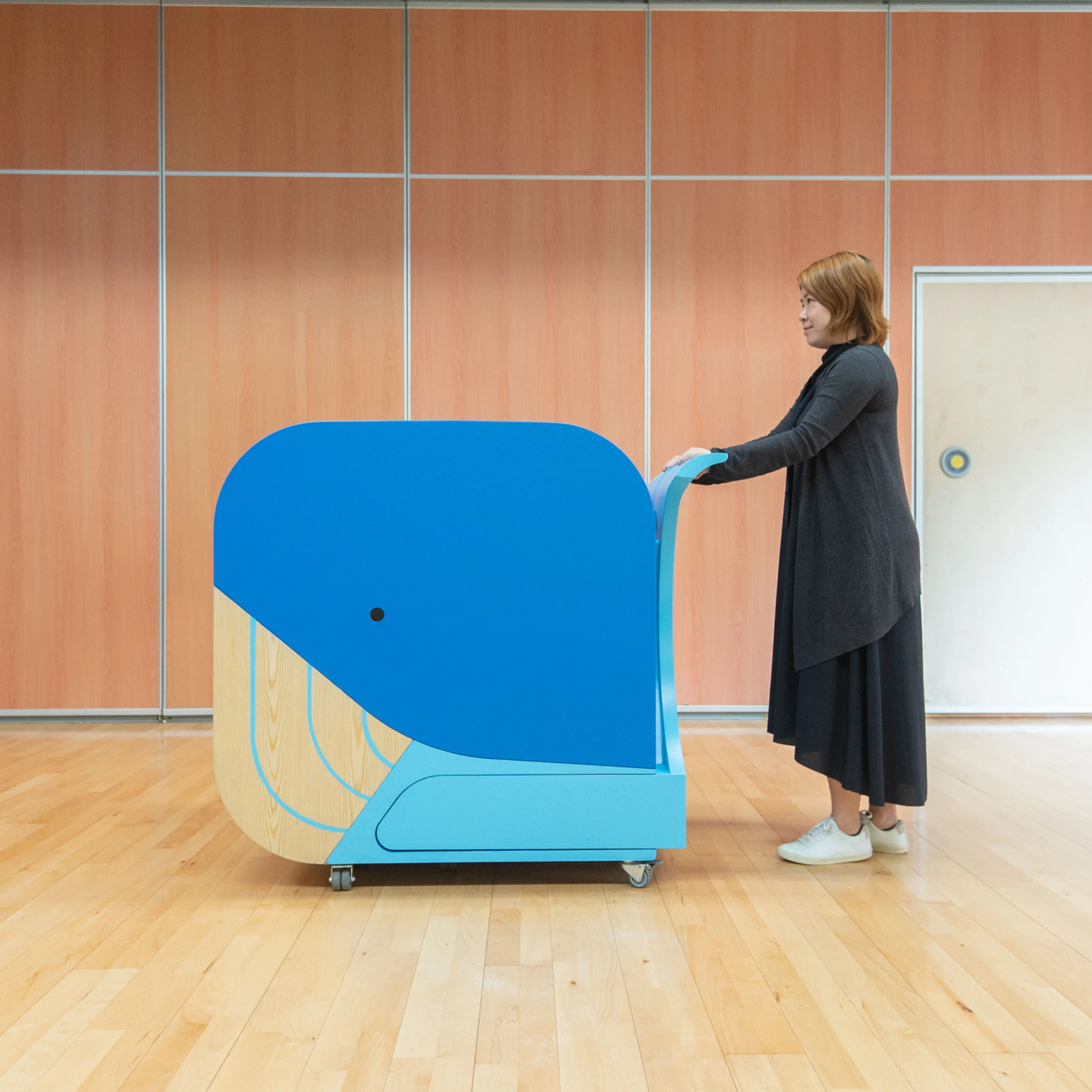 A person pushes a mobile, blue whale-shaped library cart on casters across a wood floor by HIR Studio.