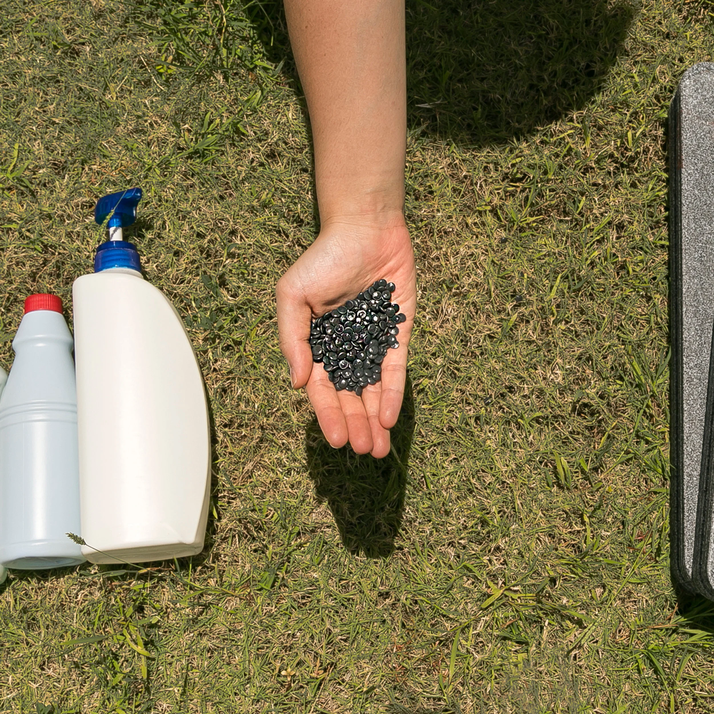 Close-up of a hand holding dark recycled plastic pellets alongside plastic bottles on grass by HIR Studio.