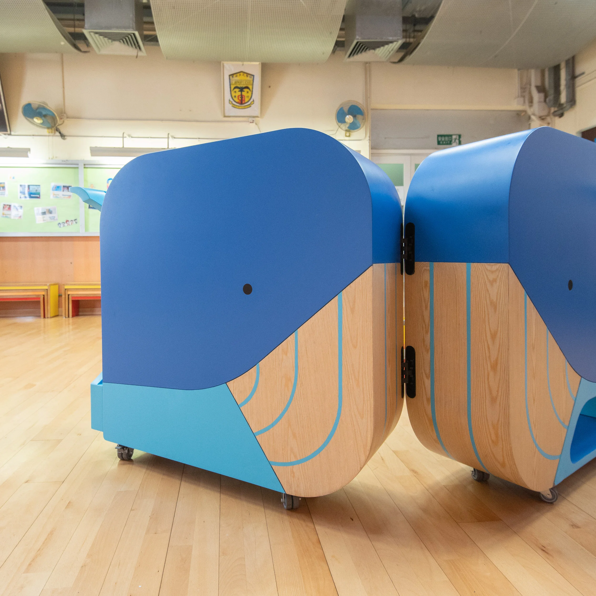 Two mobile whale-shaped modular library carts in wood and blue finish by HIR Studio inside an interior space.