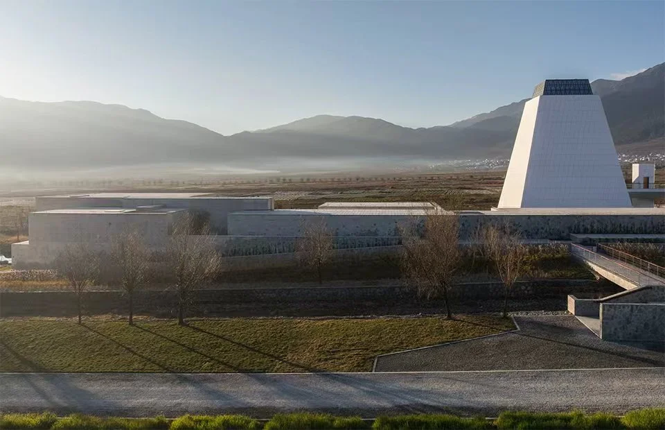 Exterior view of OLI Architecture project with low horizontal stone volumes and a prominent pyramidal tower against a mountainous backdrop.