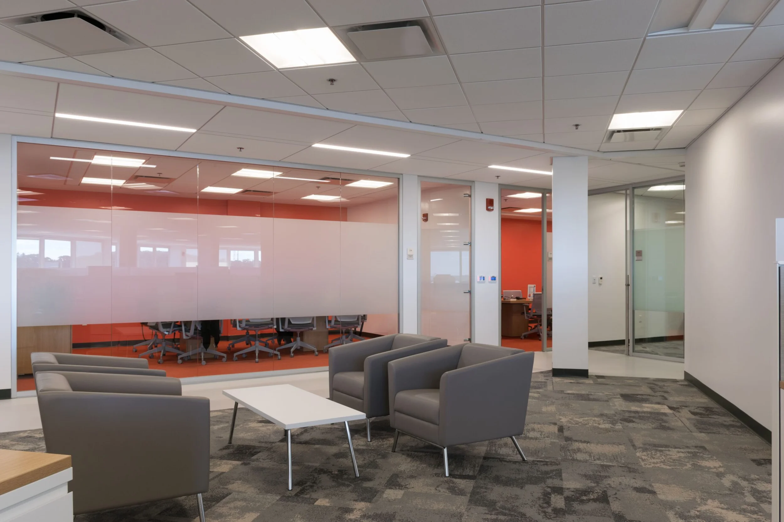 Office reception lounge with grey armchairs, a low white coffee table, patterned carpet, and glass meeting rooms with orange walls by Jarmel Kizel Architects.