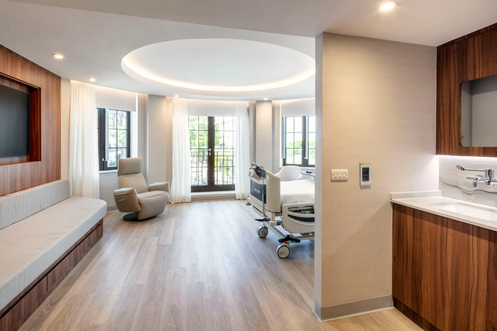 Private hospital recovery suite with curved illuminated ceiling recess, wood floors, and warm cabinetry by LSI Architects.