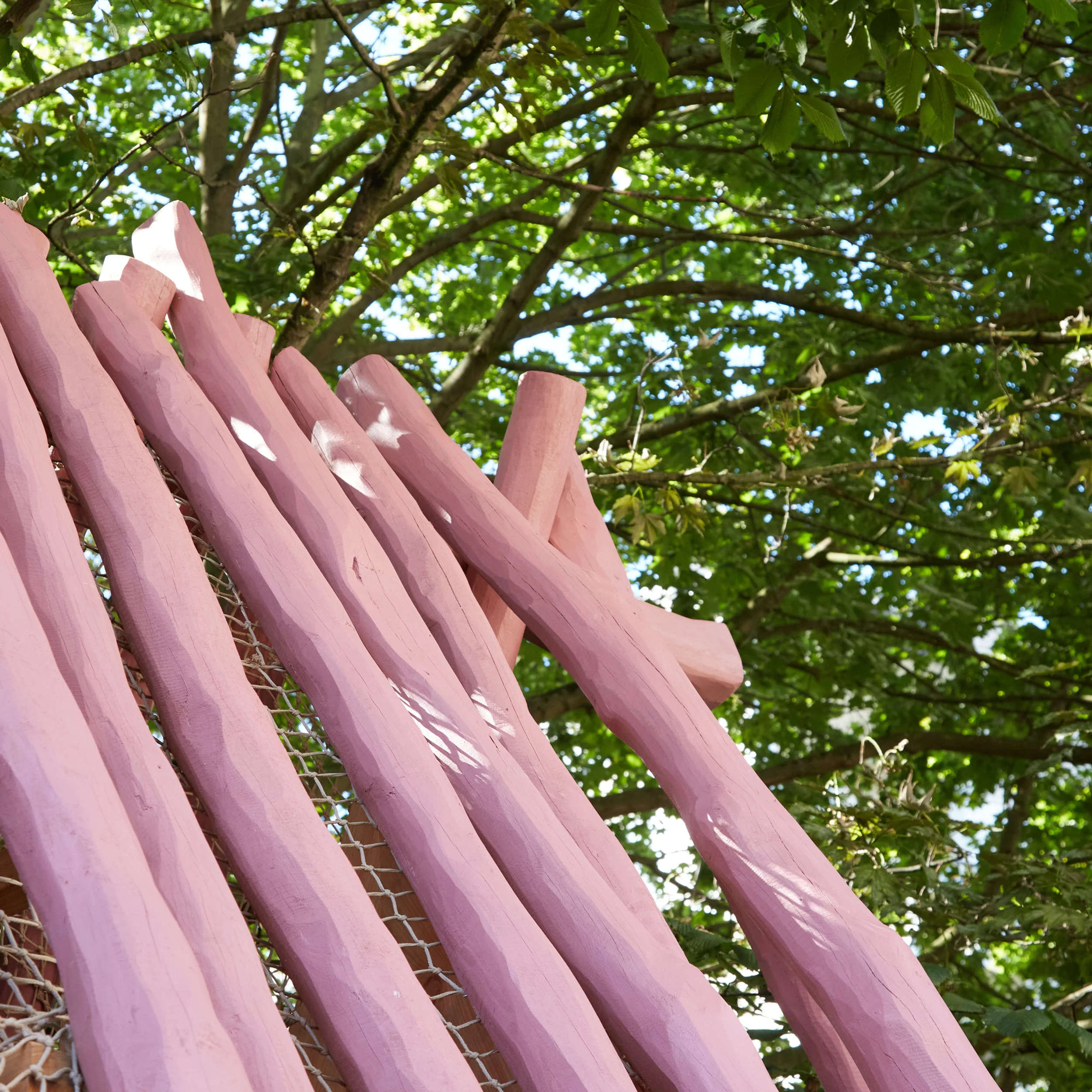 Low-angle view of pink-painted rounded log structure against a leafy green tree canopy by House of Kin.