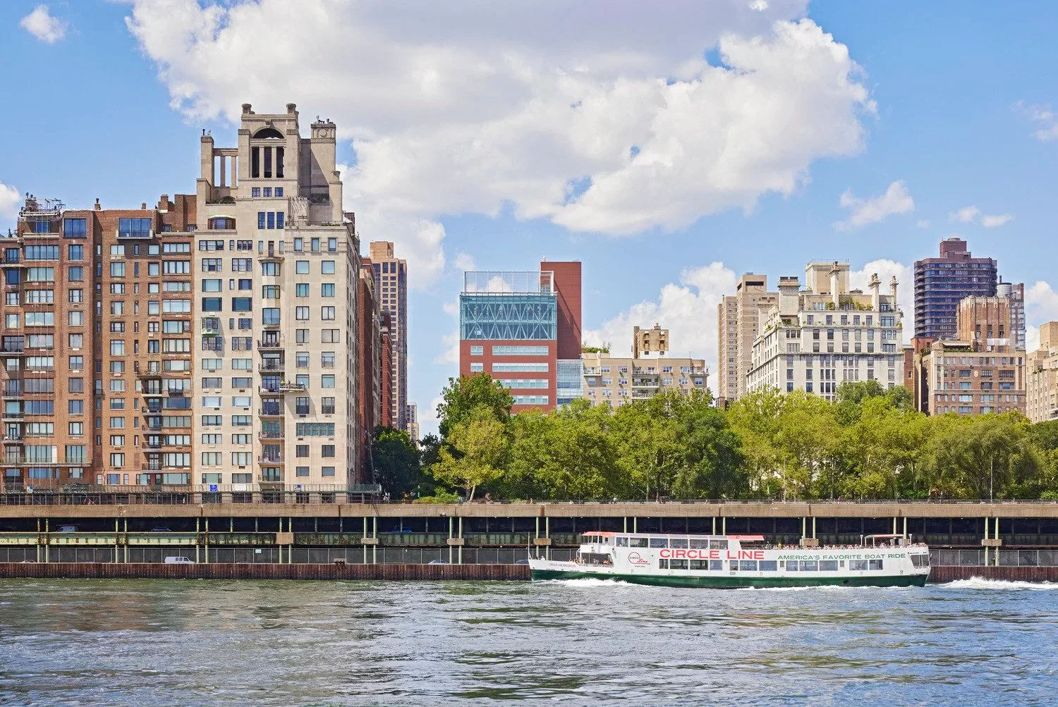 Waterfront view of an urban river edge, featuring historic and modern apartment towers behind a concrete esplanade and a passing tour boat.