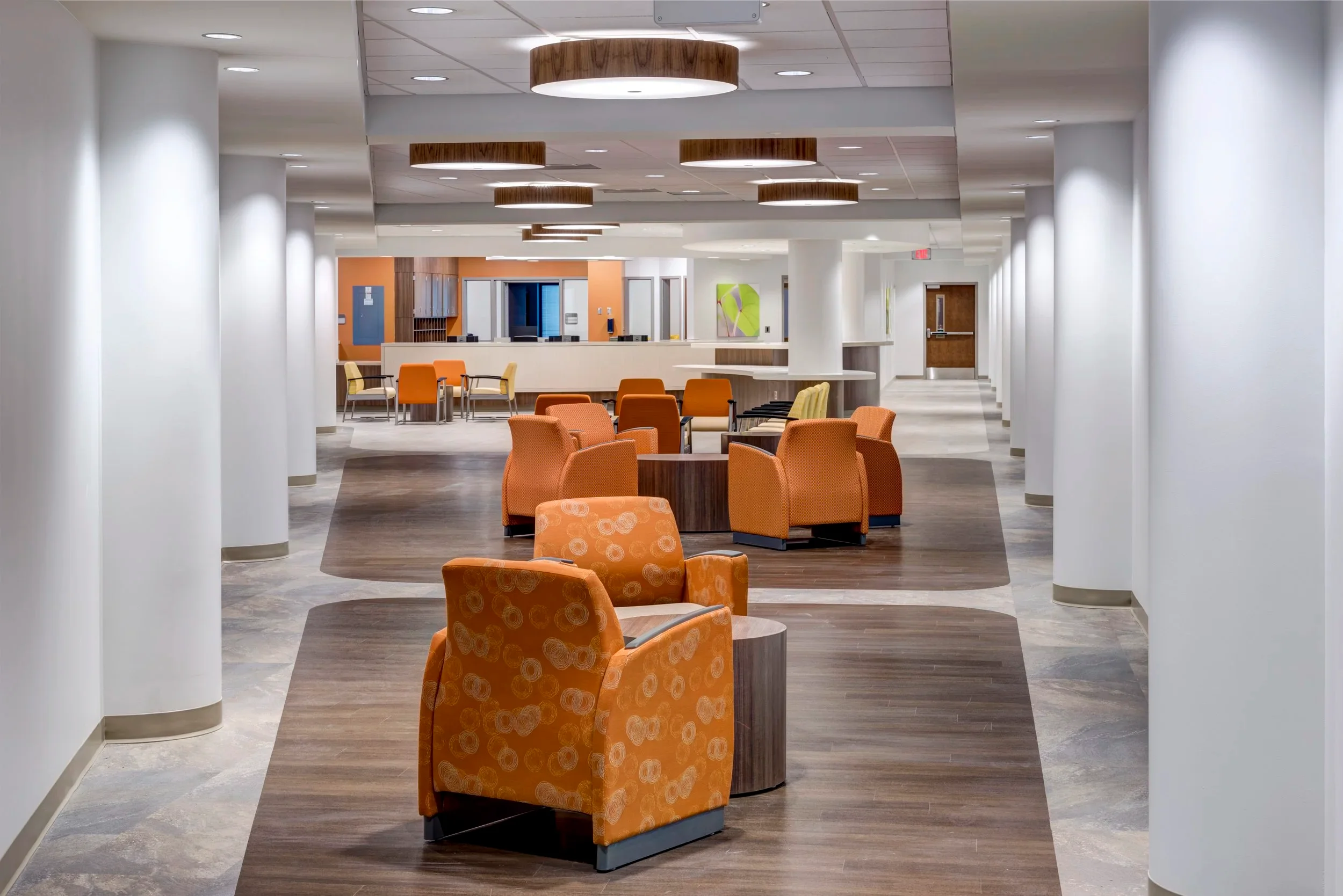 Spacious healthcare interior corridor by NK Architects featuring round white columns, orange armchairs, and wood-inlaid flooring areas.