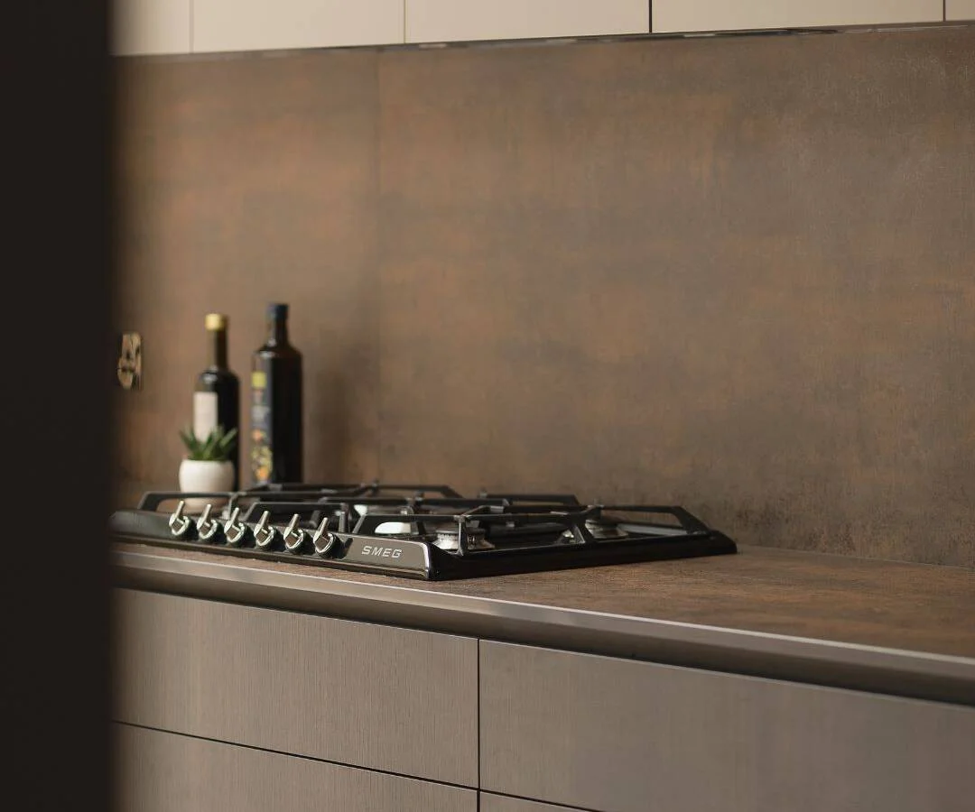 Close-up of a kitchen cooktop set against a rusted metal backsplash and dark cabinetry by Pencil and Brick.