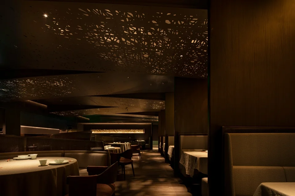Dark interior of a restaurant featuring intricate perforated ceiling panels casting dappled light patterns by SIG STUDIO.