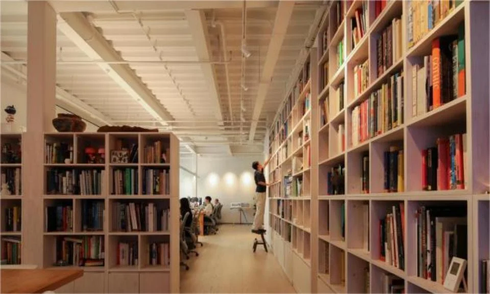 Interior of an office space by SIG STUDIO featuring extensive floor-to-ceiling white bookshelf walls and exposed ribbed ceiling.