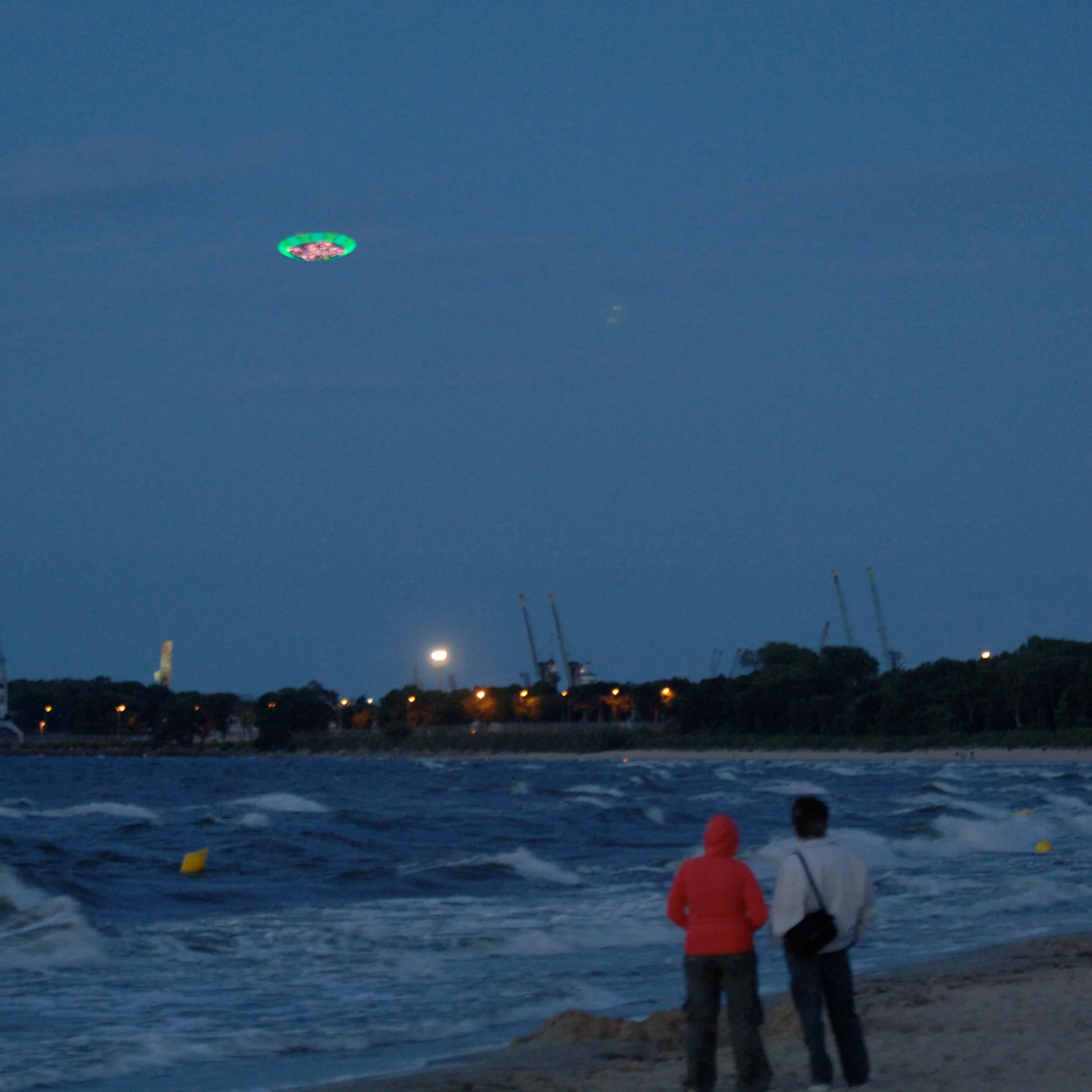 Two people on a sandy beach at dusk watching a glowing green art installation hovering over the dark water in Gdansk by Cinimod Studio.