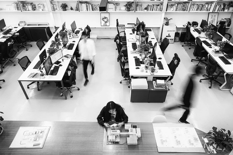 High-angle view of a busy architecture studio workspace by 城外建筑 in Guangzhou, featuring long work tables, bookshelves, and blurred figures in motion.