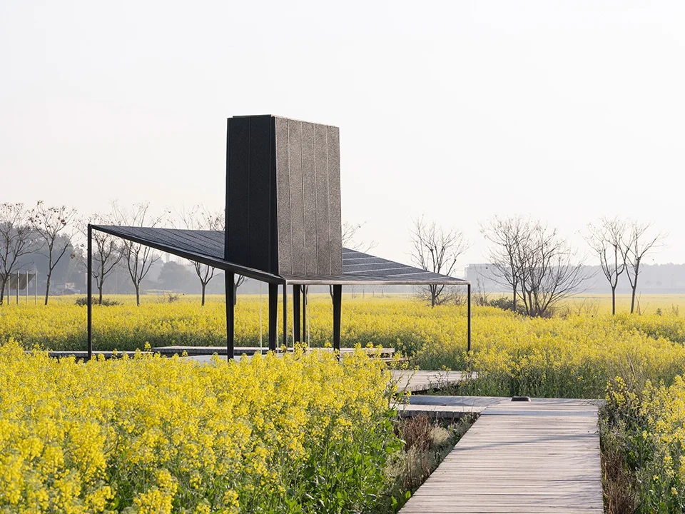 A black pavilion with a tall vertical volume and angled roof floating over a yellow field with timber boardwalks.
