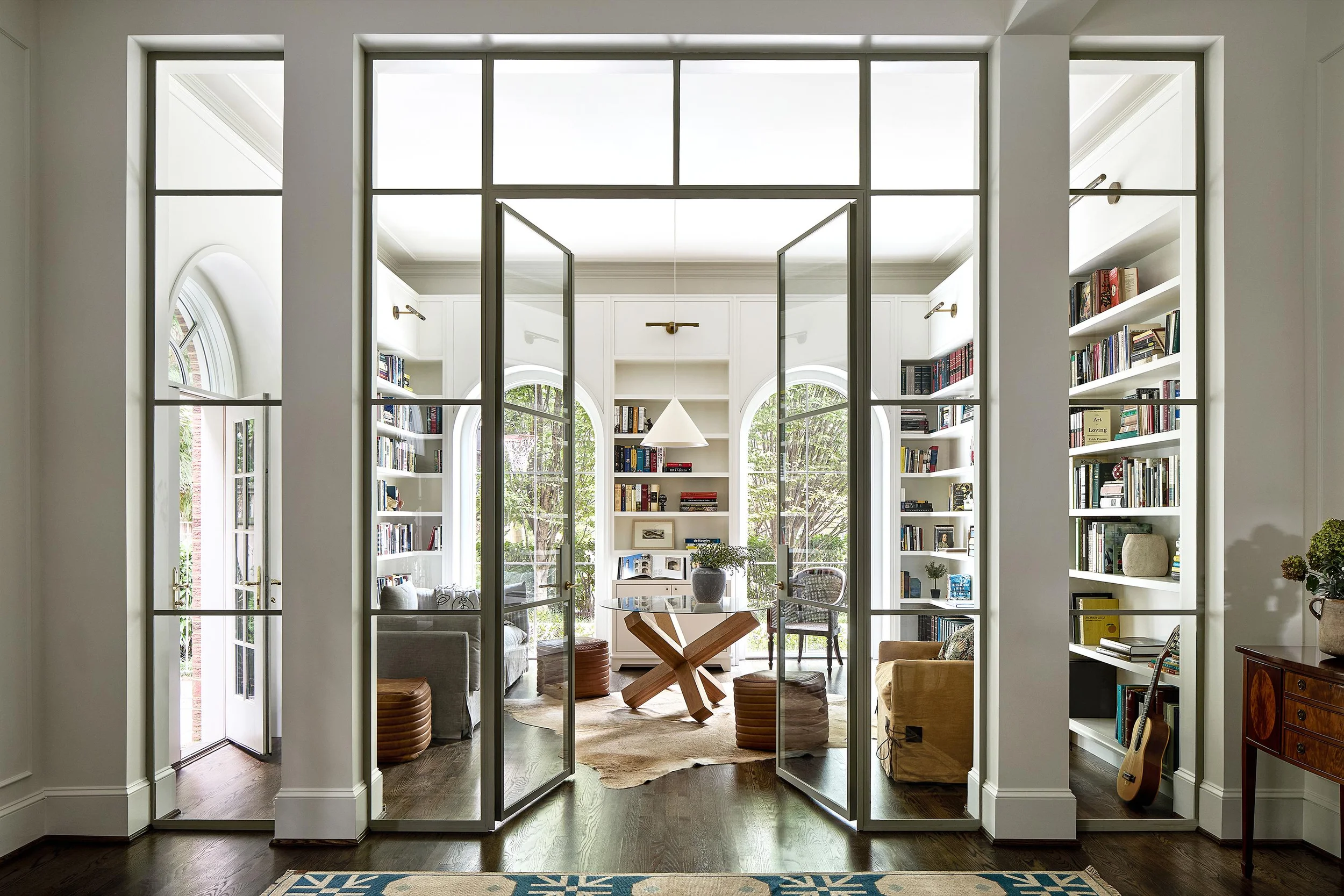 Interior view through slender steel-framed glass double doors into a symmetrical book-lined study by Fowlkes Studio.