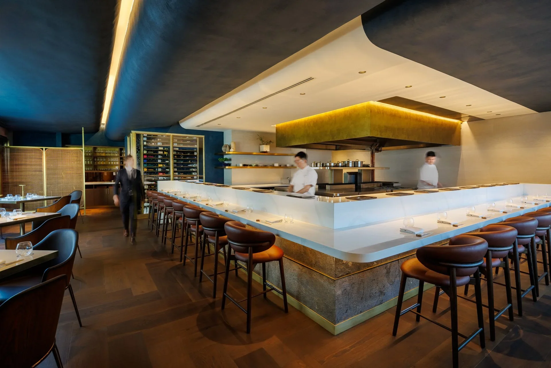 Sushi bar interior with a wrap-around white stone counter and curved dark leather stools by Studio UNLTD.