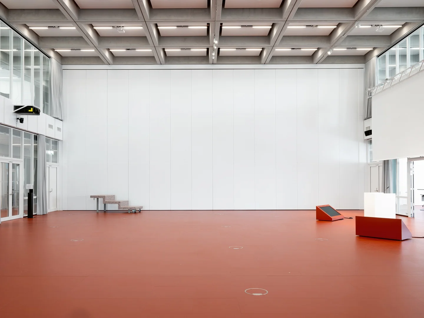 Interior of a large hall by Christ & Gantenbein featuring a deep coffered concrete ceiling, red linoleum floor, and large white operable acoustic partition wall.