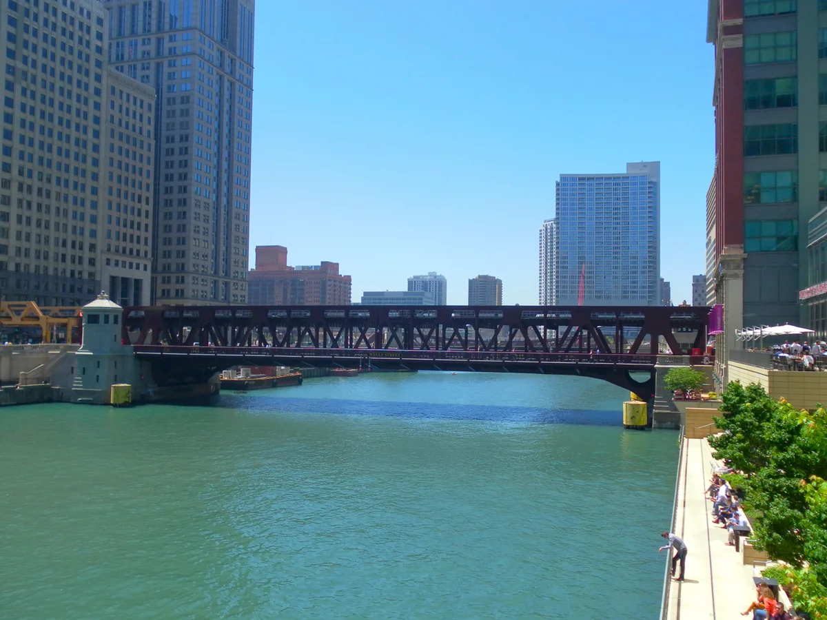 View of a historic steel truss movable bridge spanning a city river, flanked by high-rise towers and a public riverwalk.