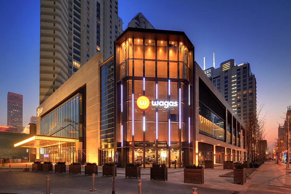 Dusk exterior view of a multi-story commercial retail podium with a glowing curved glass corner facade and vertical light strips by Cendes.